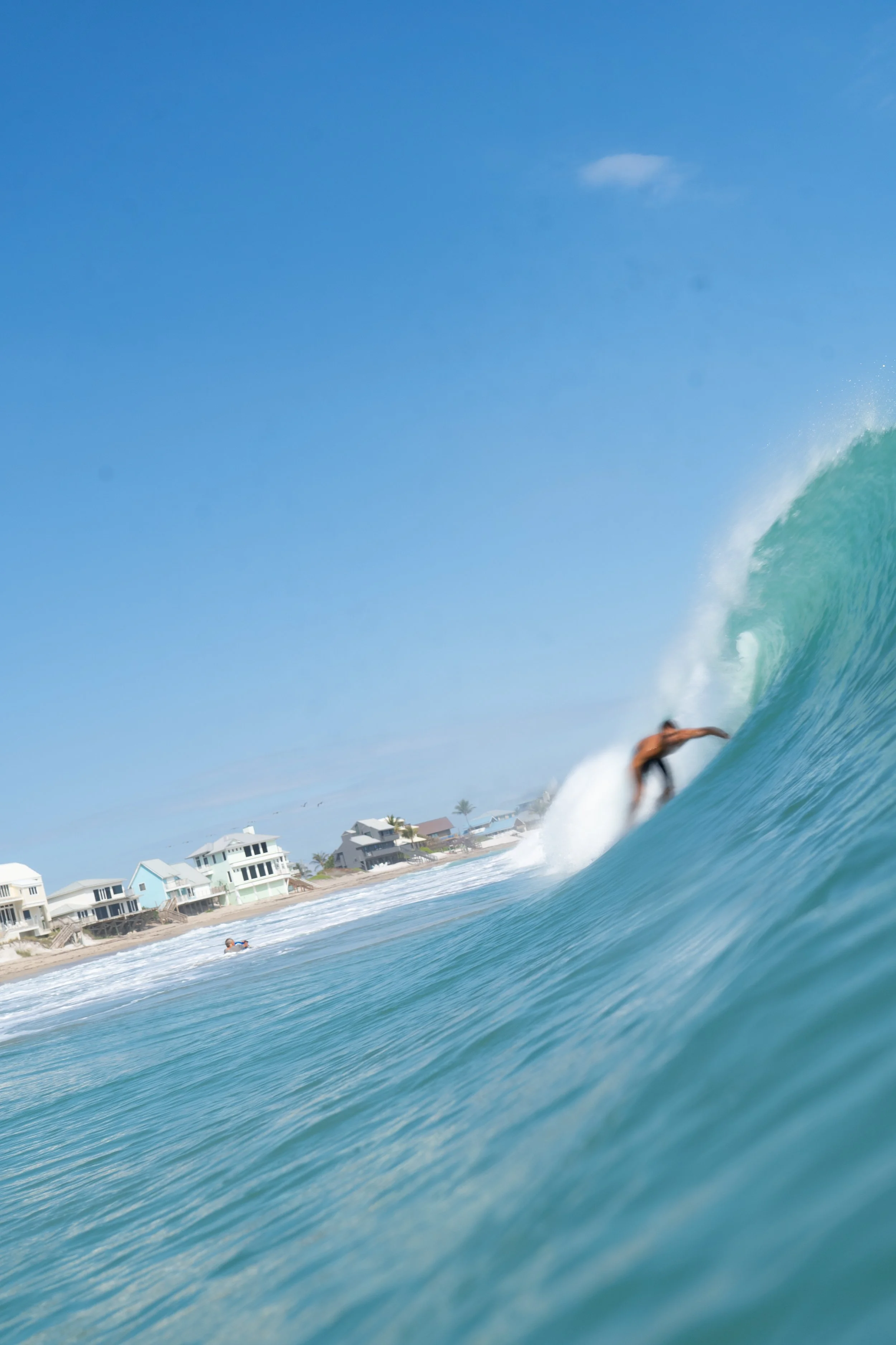 A person surfing a large wave near a coastal area with houses and palm trees in the background under a clear blue sky.