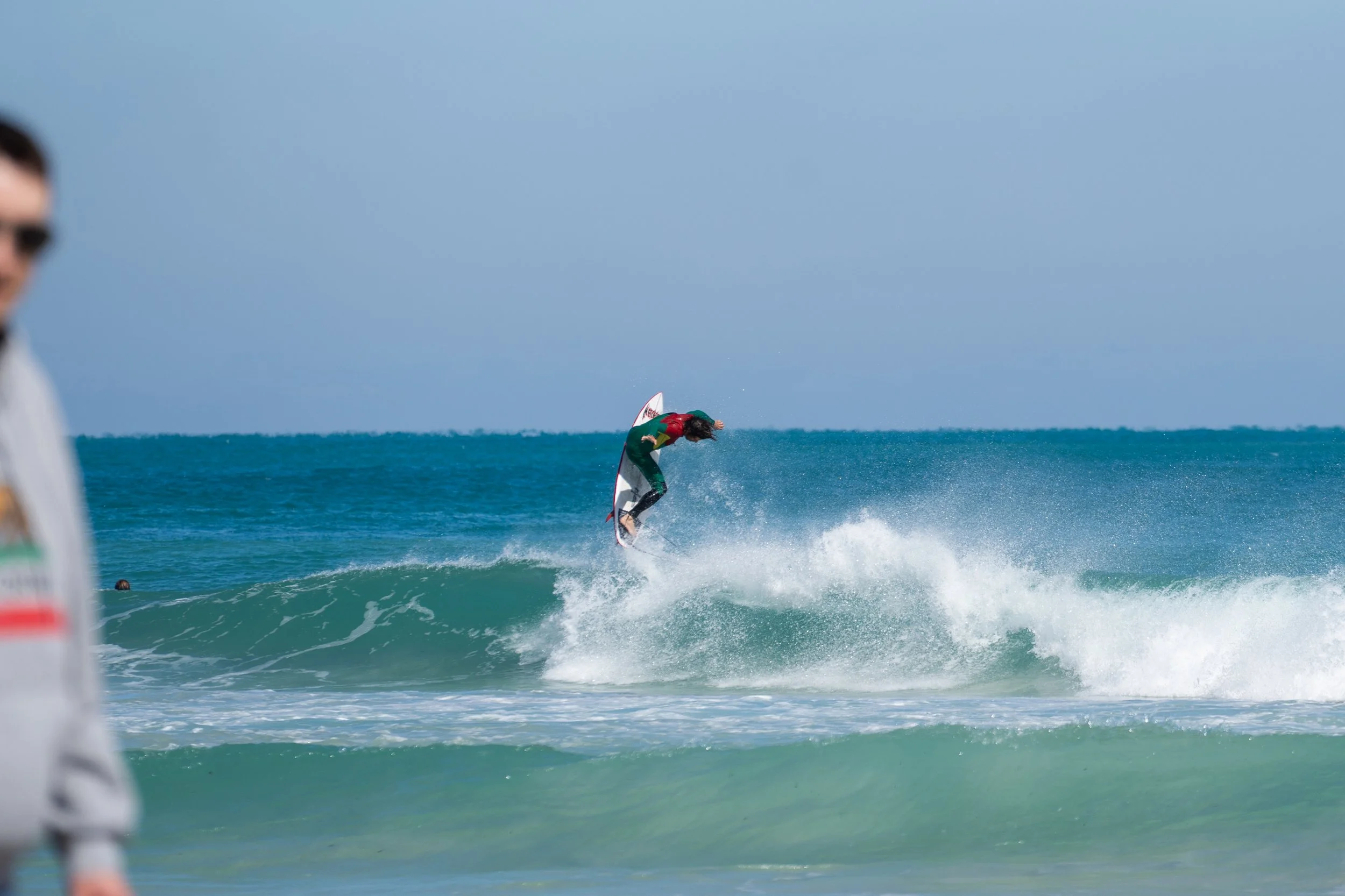 A person in a red and green wetsuit riding a wave on a surfboard in the ocean, with another person in the foreground on the left side of the image.