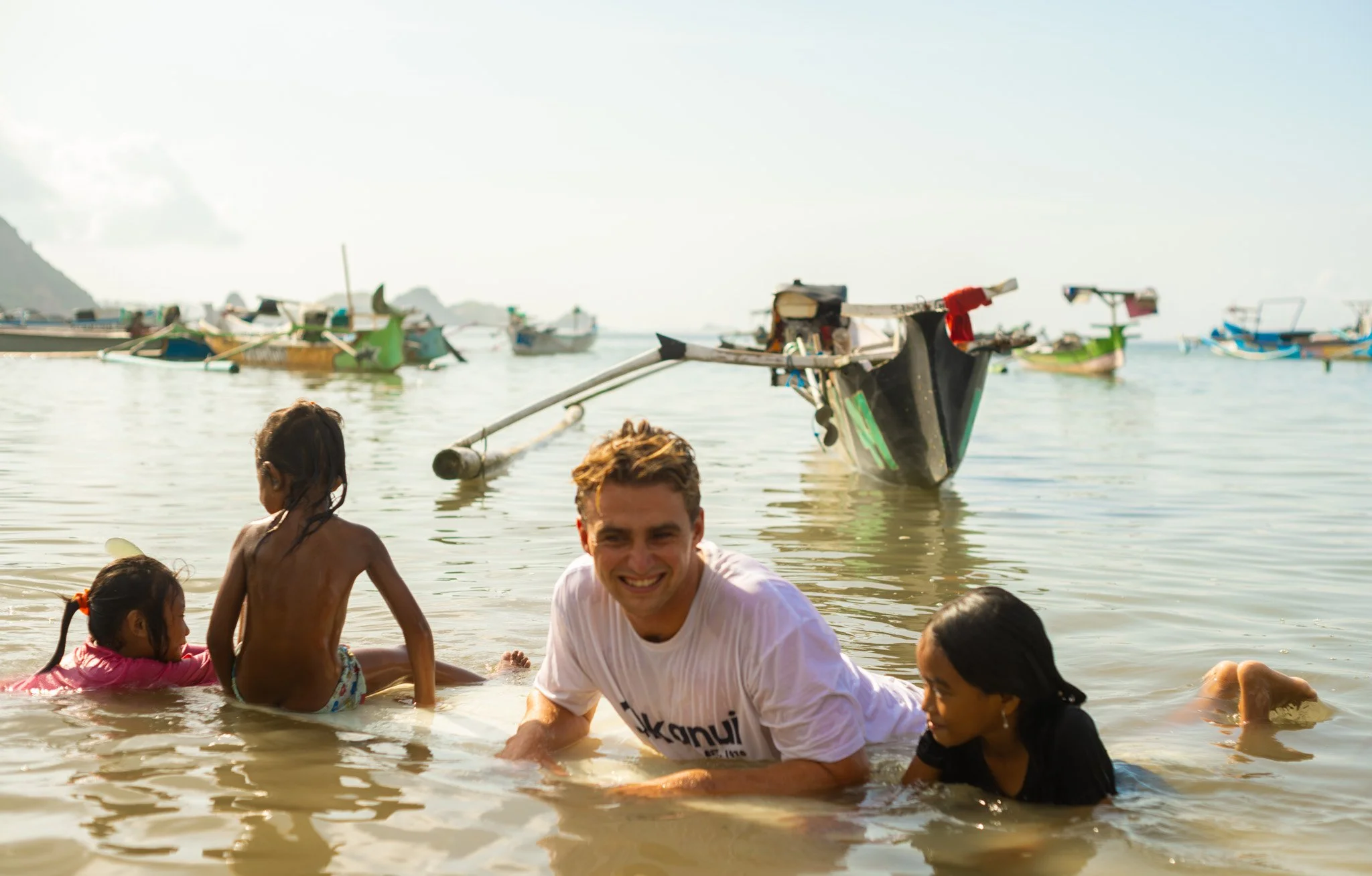 People swimming and playing in shallow water near boats on a sunny day at the beach.