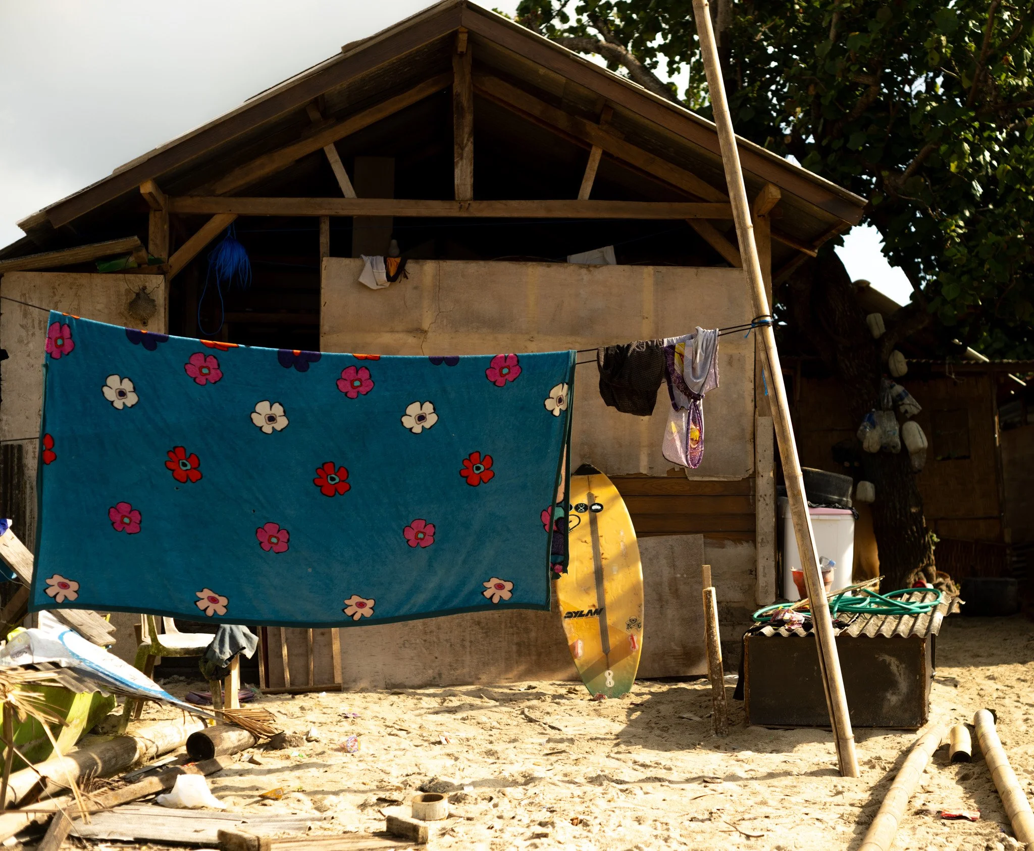 A beach scene with a homemade wooden house in the background, a colorful towel with flower patterns hanging on a clothesline, and various objects including a skateboard, a chest, and some pipes on sandy ground.