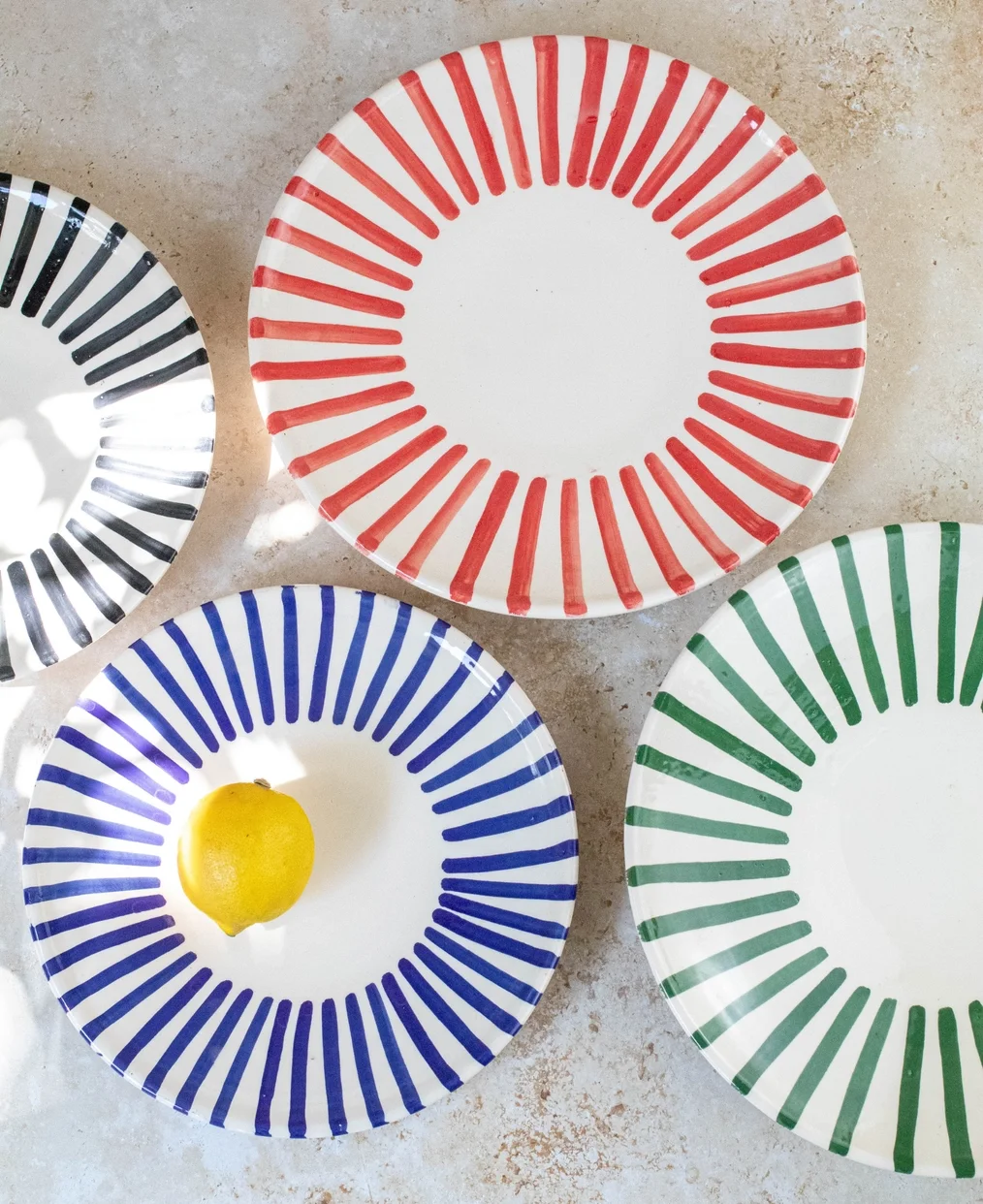 Four decorative plates with colorful radial stripe patterns in red, black, blue, and green on a beige surface, one plate with a lemon on it.