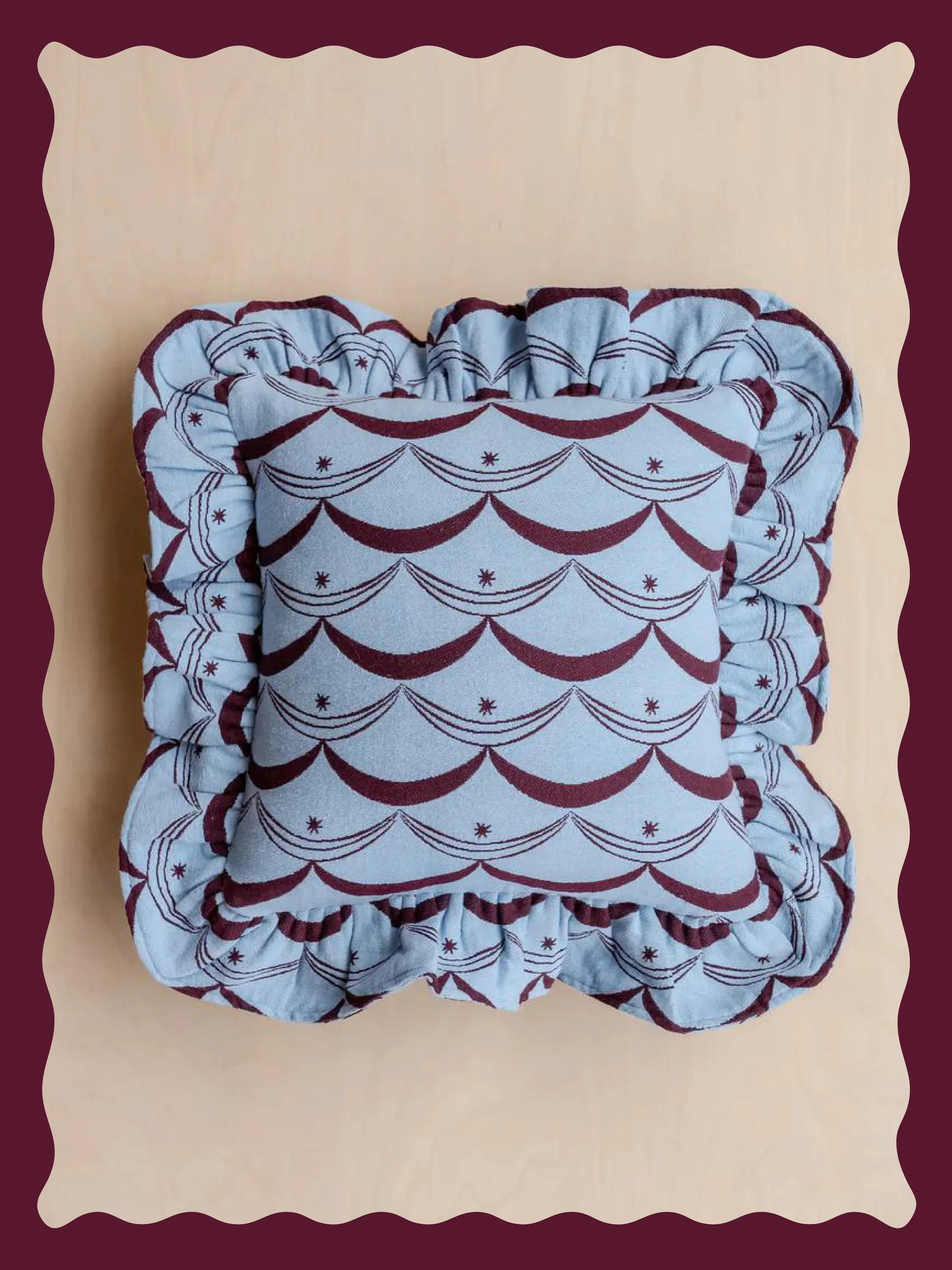 Stacked square cushions with ruffled edges, featuring a pattern of maroon, lace, and star motifs on a light blue background.