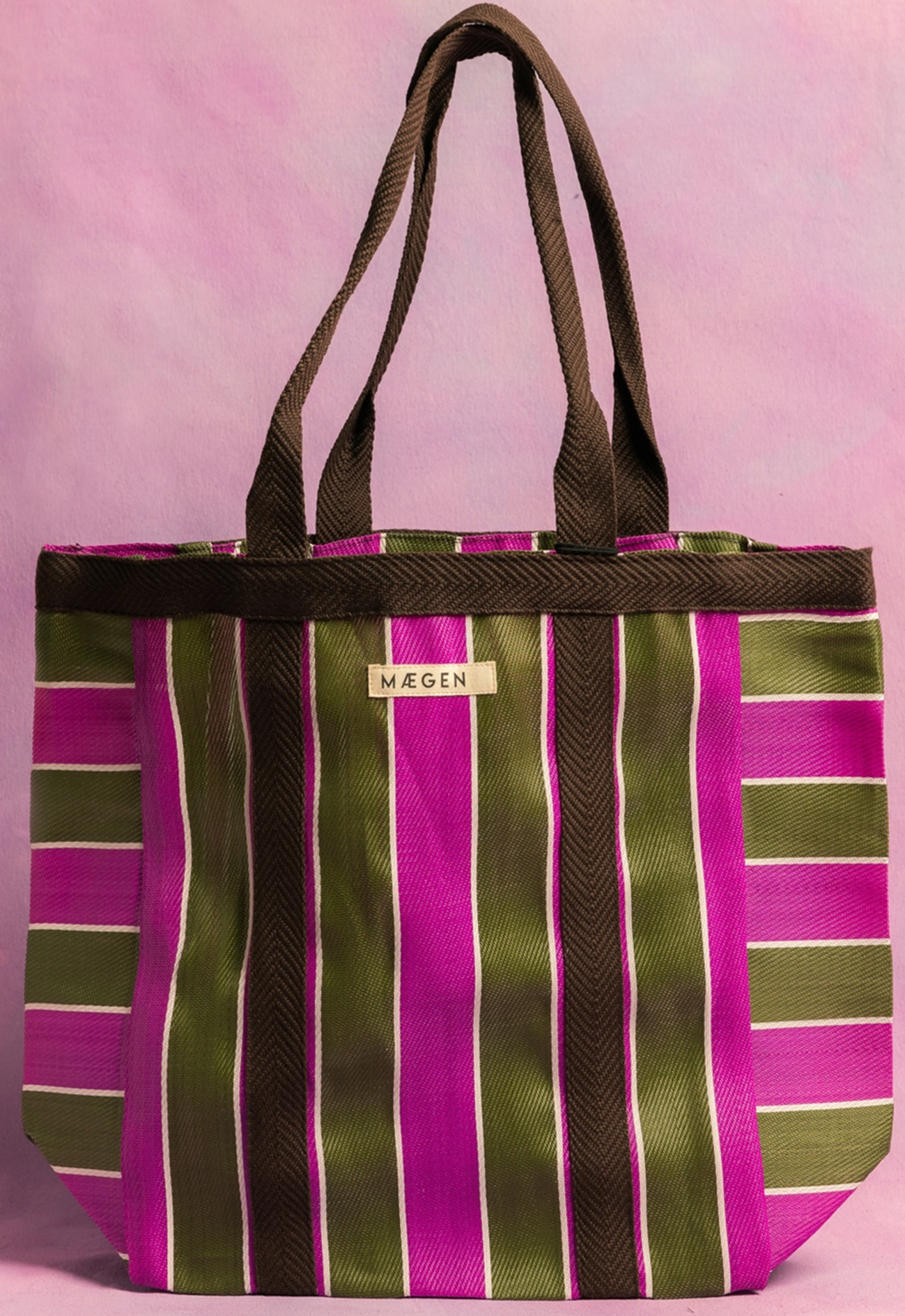 A striped tote bag with pink, green, and brown vertical stripes, brown handles, and a label that says 'MAEGEN' on the front, set against a pink background.