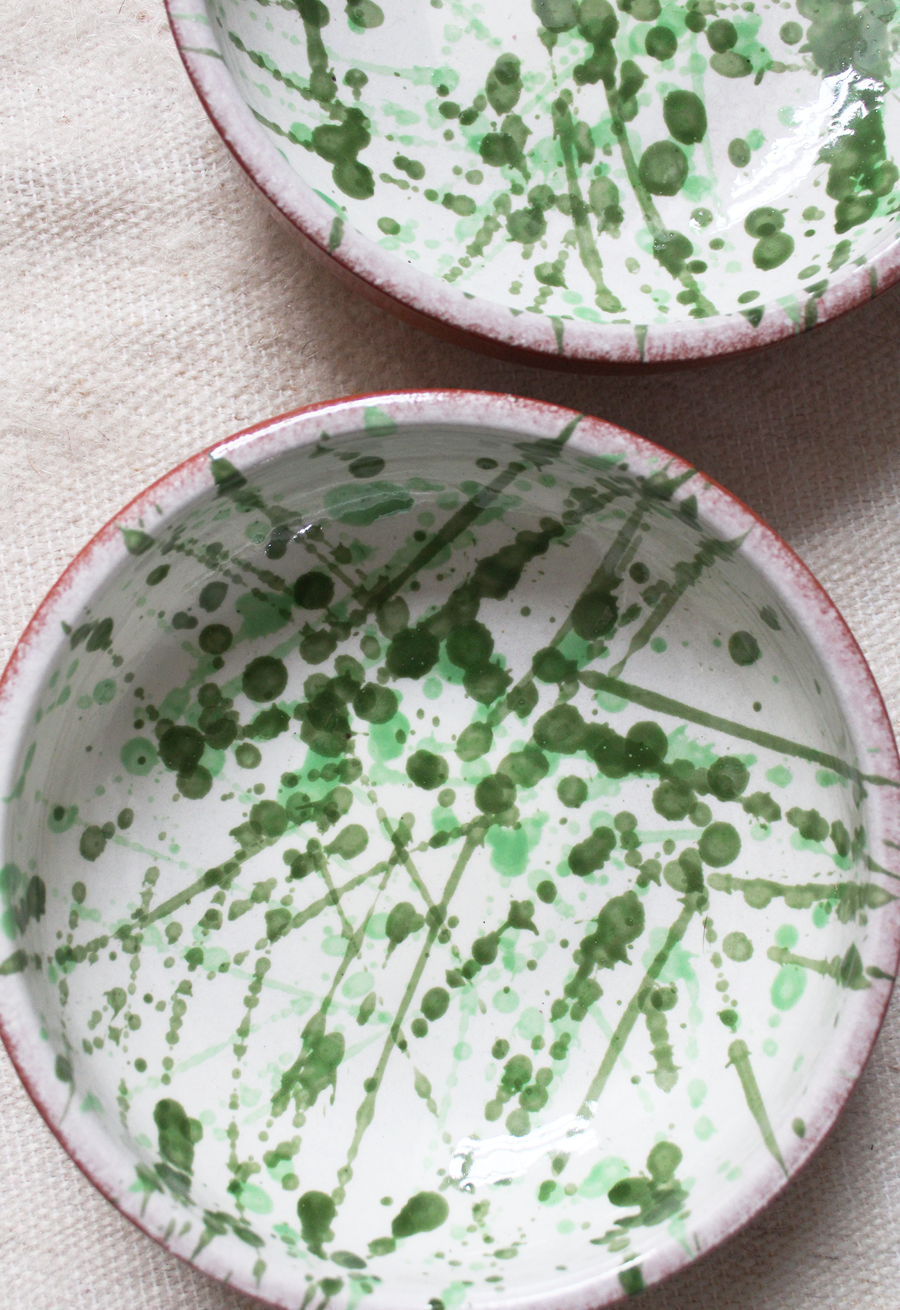 Two ceramic bowls with green splatter paint designs on the interior, placed on a beige fabric surface.