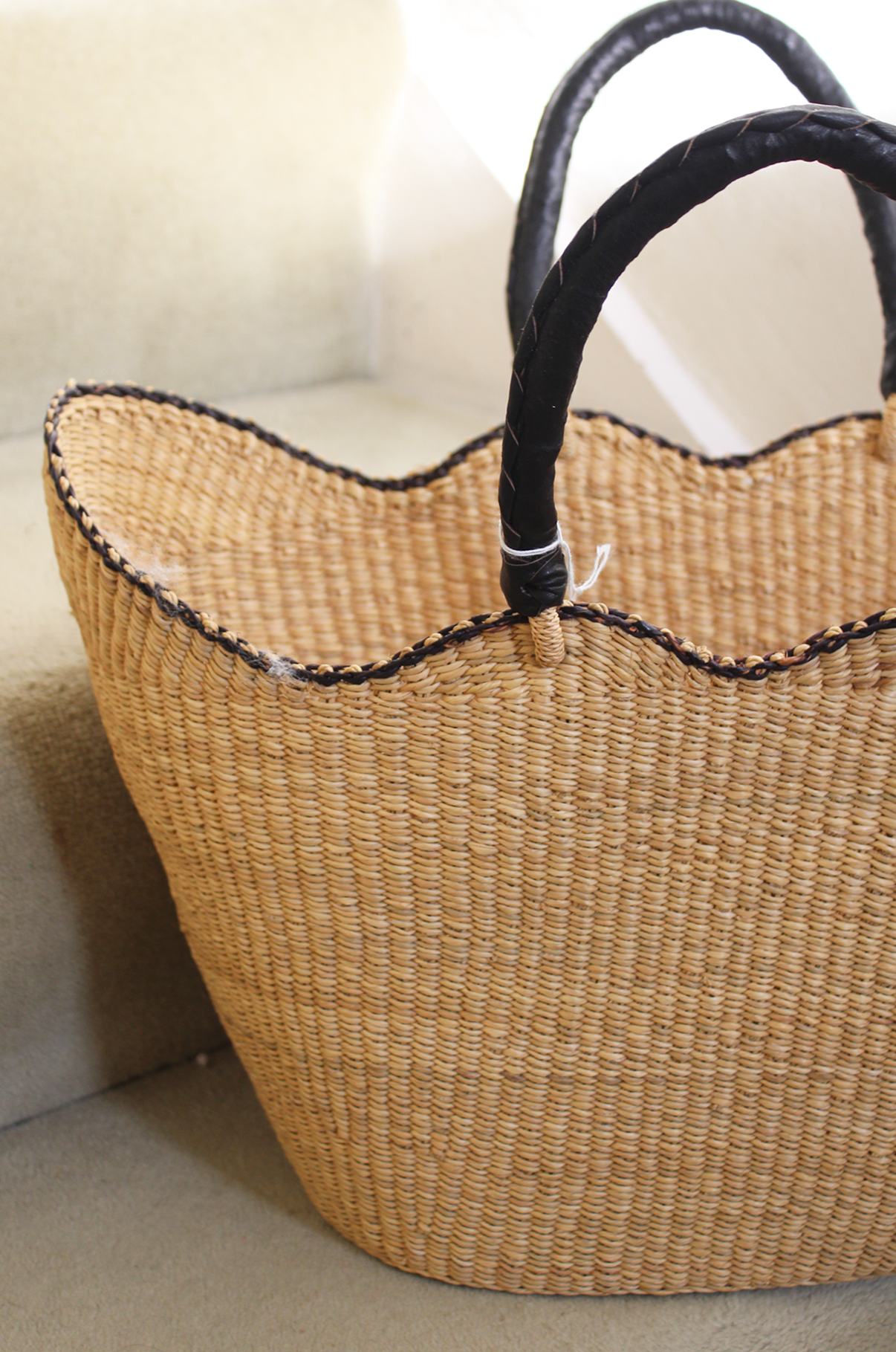 A woven straw handbag with dark leather handles resting on a beige surface.