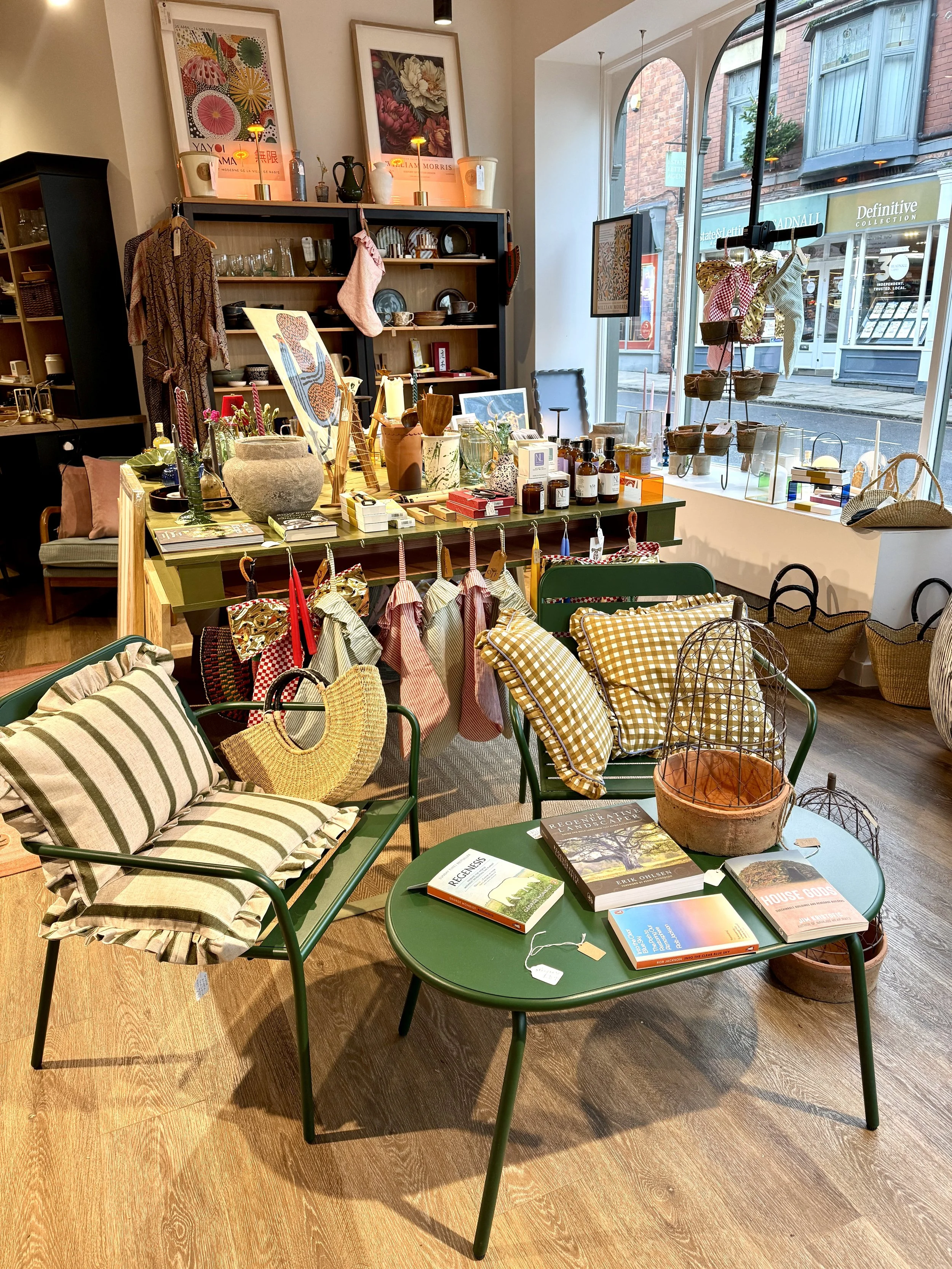A cozy boutique with a variety of home decor, furniture, and gifts displayed, including pillows, books, vases, and intricate baskets, with large windows letting in natural light.