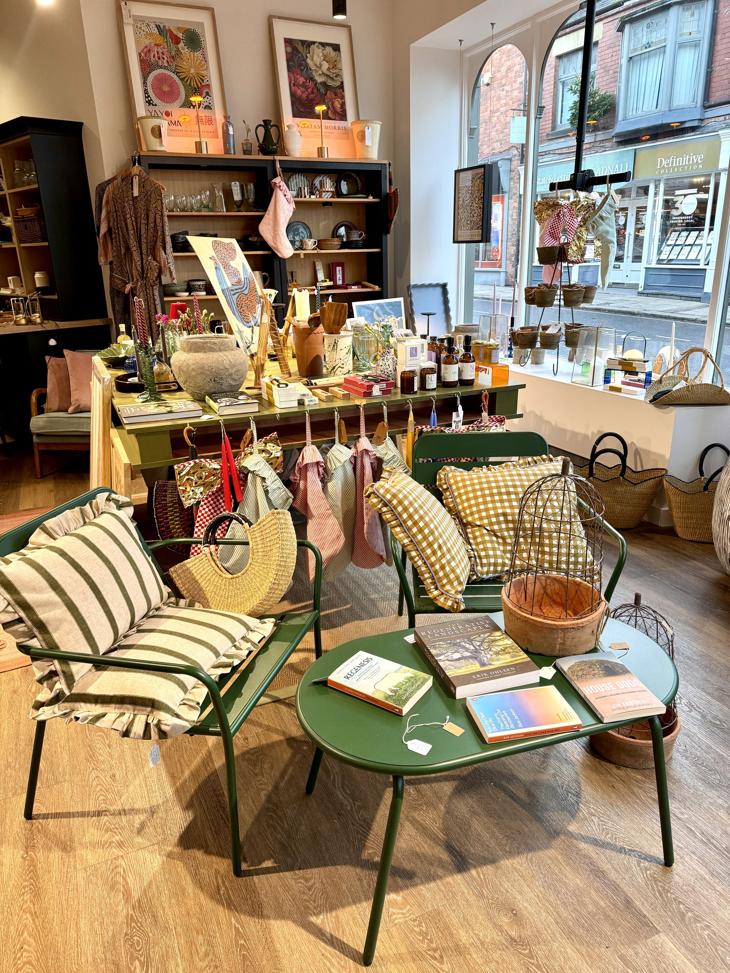 Interior of a boutique shop with display tables, shelves, and chairs, showcasing home decor, books, and accessories near a large window.