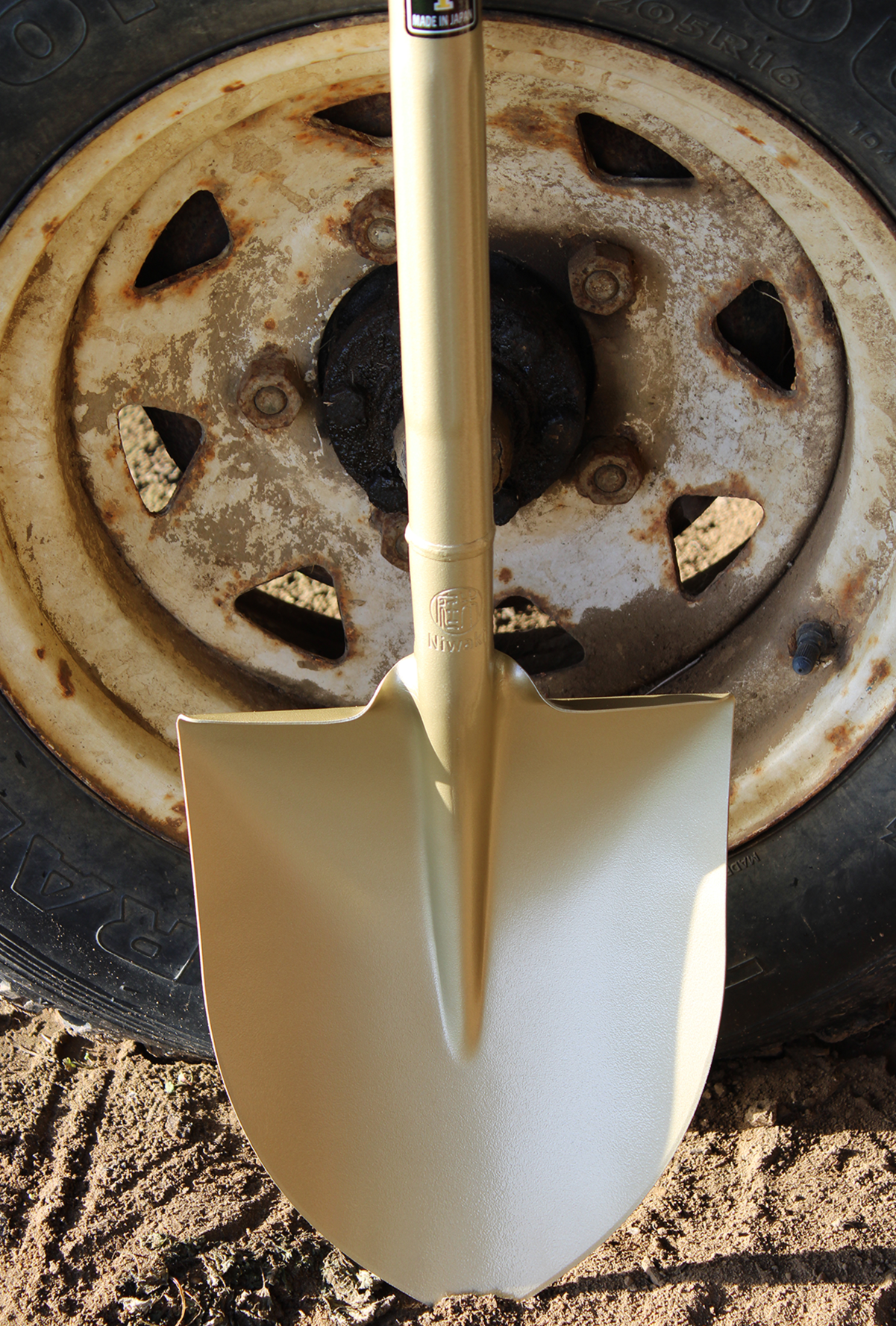 Close-up of a shiny gold shovel placed over a rusty wheel on a vehicle, with dirt and soil on the ground.