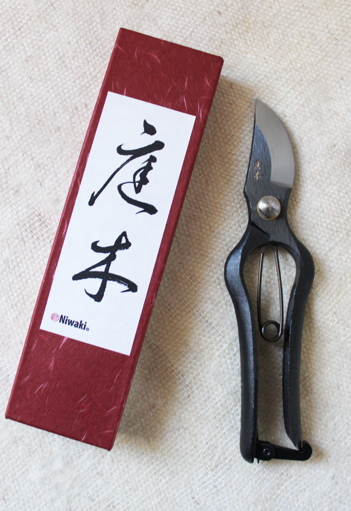 A Japanese kiridashi knife with a black handle and a short, curved blade next to a red box with Japanese characters on a textured beige surface.