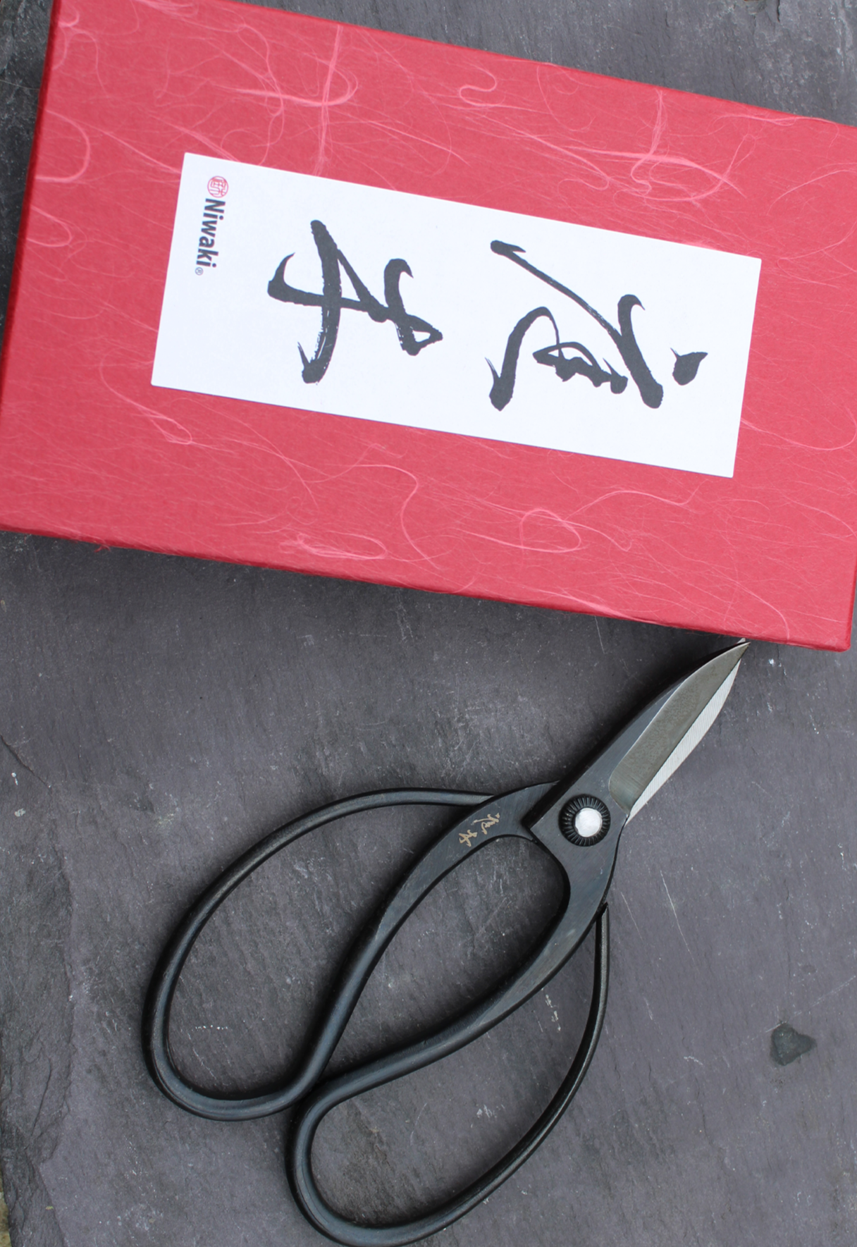 A red box with a white label and black Japanese calligraphy, positioned next to a pair of black scissors on a dark surface.