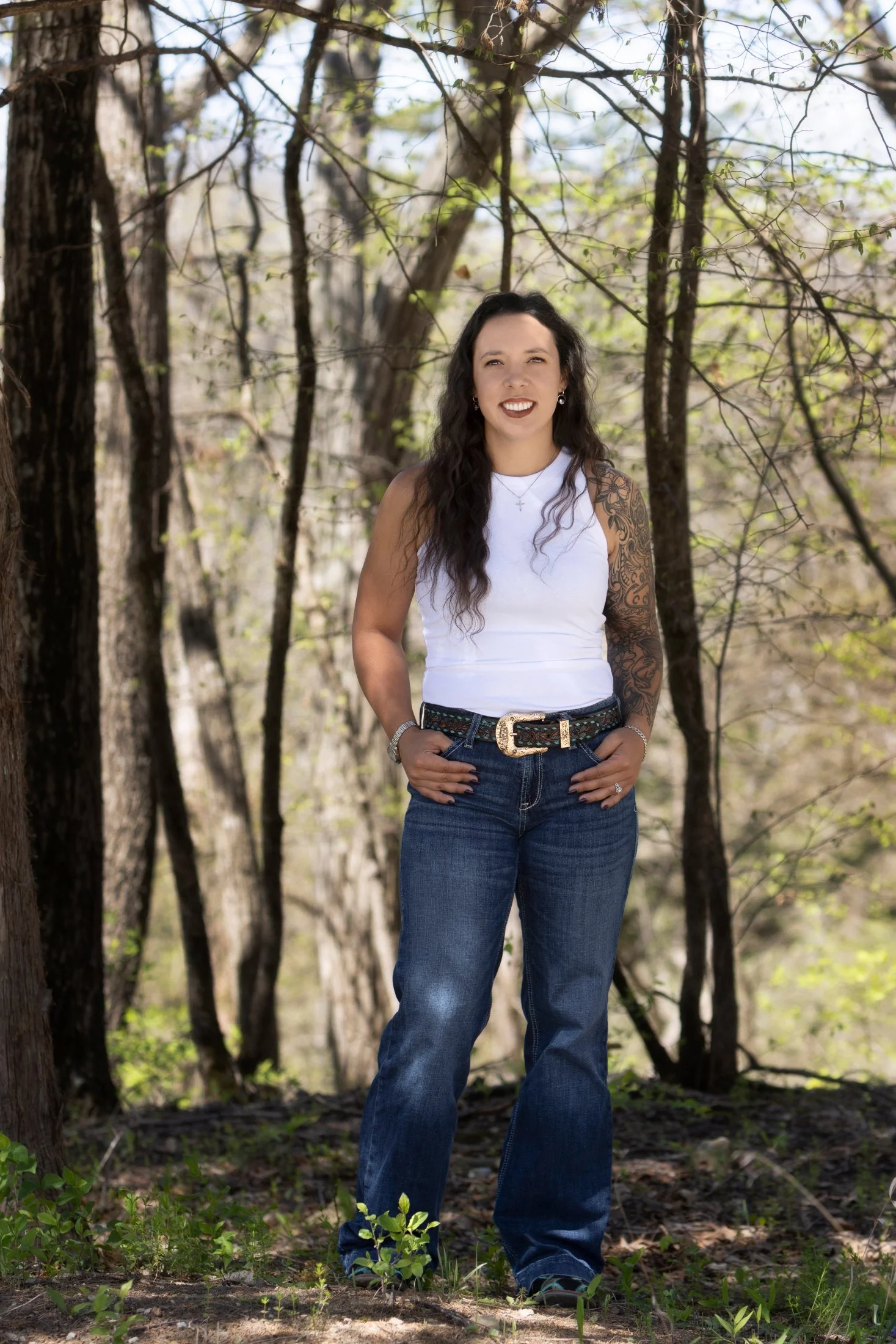 A woman with long dark hair, tattoos on her left arm, wearing a white sleeveless top, jeans, and belt, standing outdoors in a wooded area with green trees and sunlight filtering through the branches.