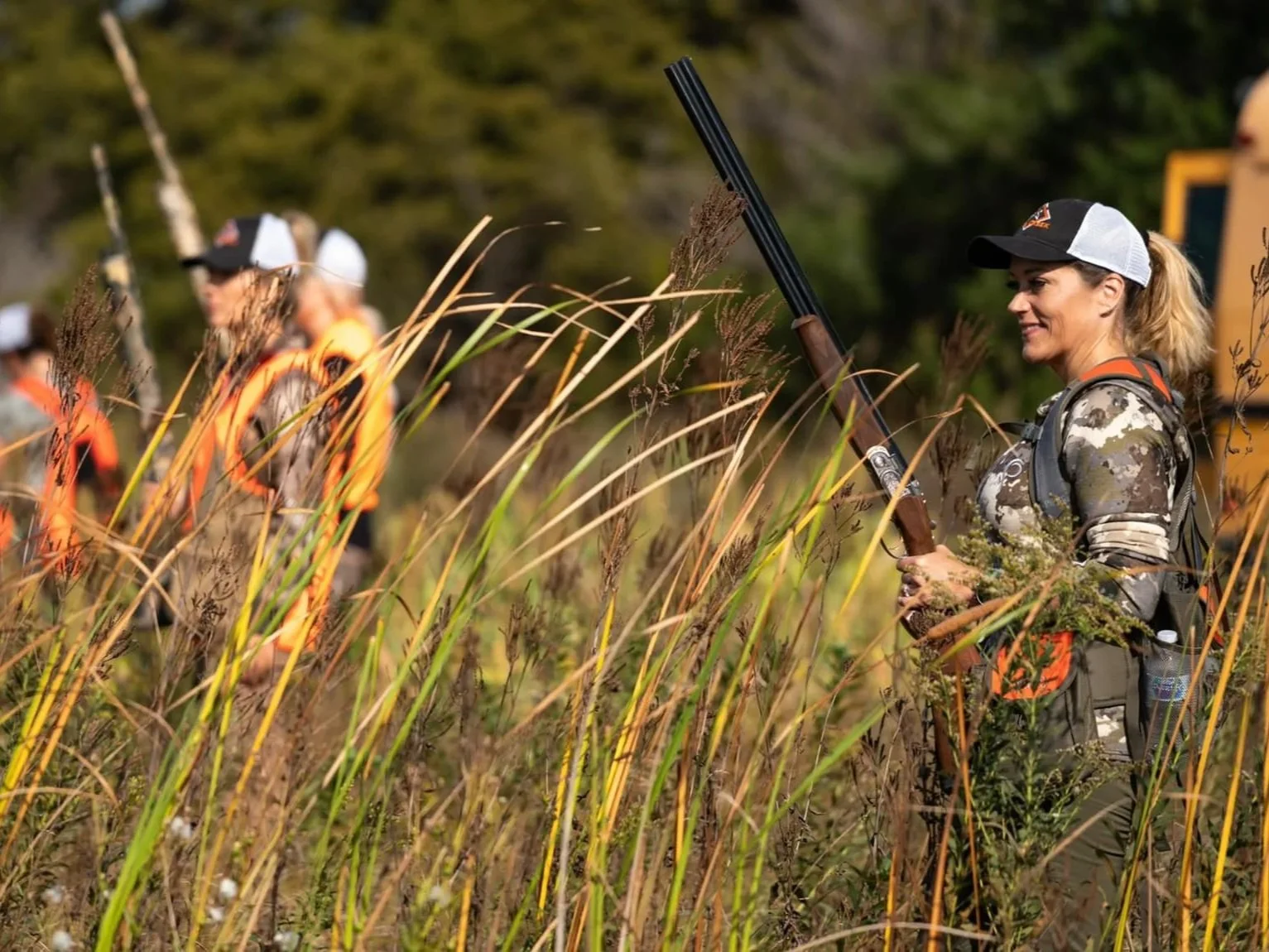 A group of hunters in camouflage and orange vests with hats, walking through tall grass in a natural outdoor area, with a female hunter holding a shotgun in the foreground.