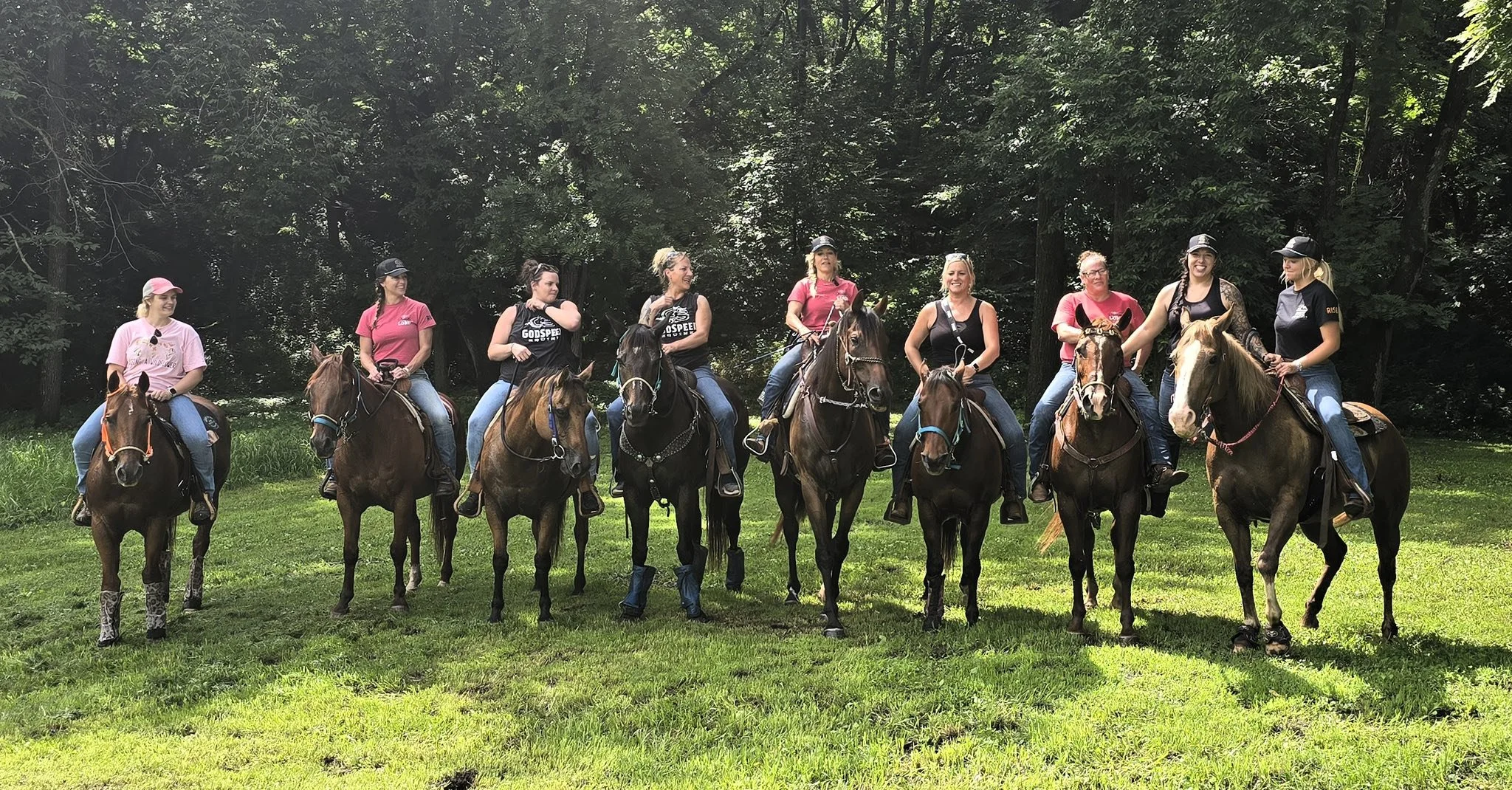 Group of women riding horses in a wooded outdoor area with green grass, wearing casual attire and riding helmets.