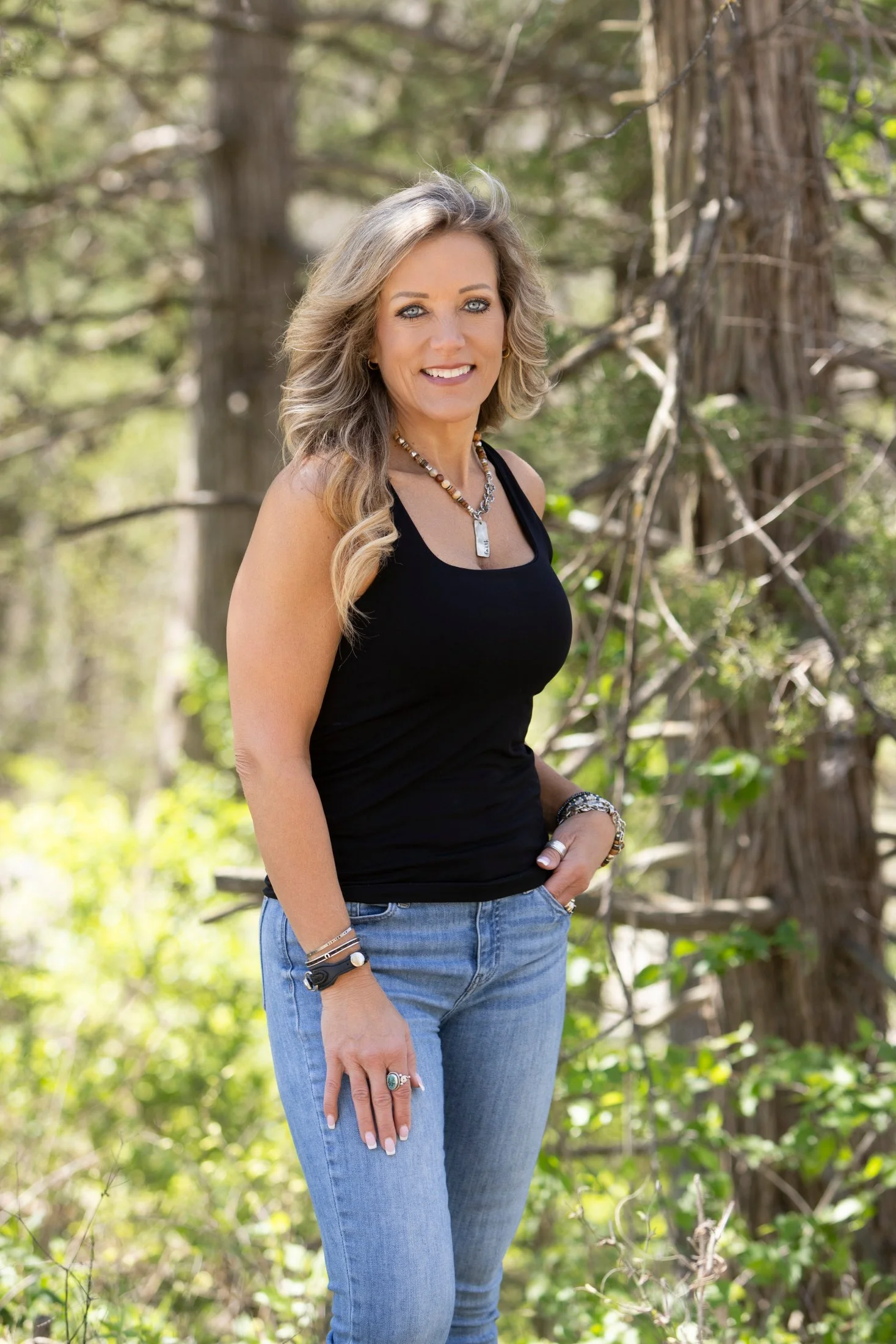 A woman with gray hair wearing a black tank top and blue jeans, standing outdoors in a wooded area with trees and green foliage.