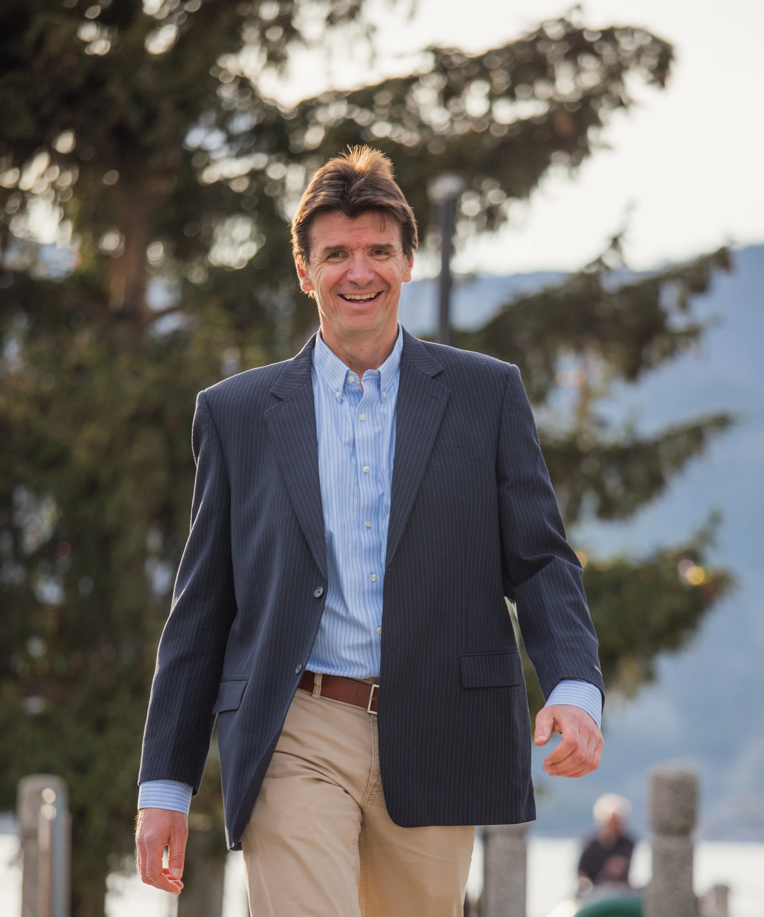 A man dressed in a blue blazer, light blue dress shirt, and tan pants walking outdoors with trees and a mountain in the background.