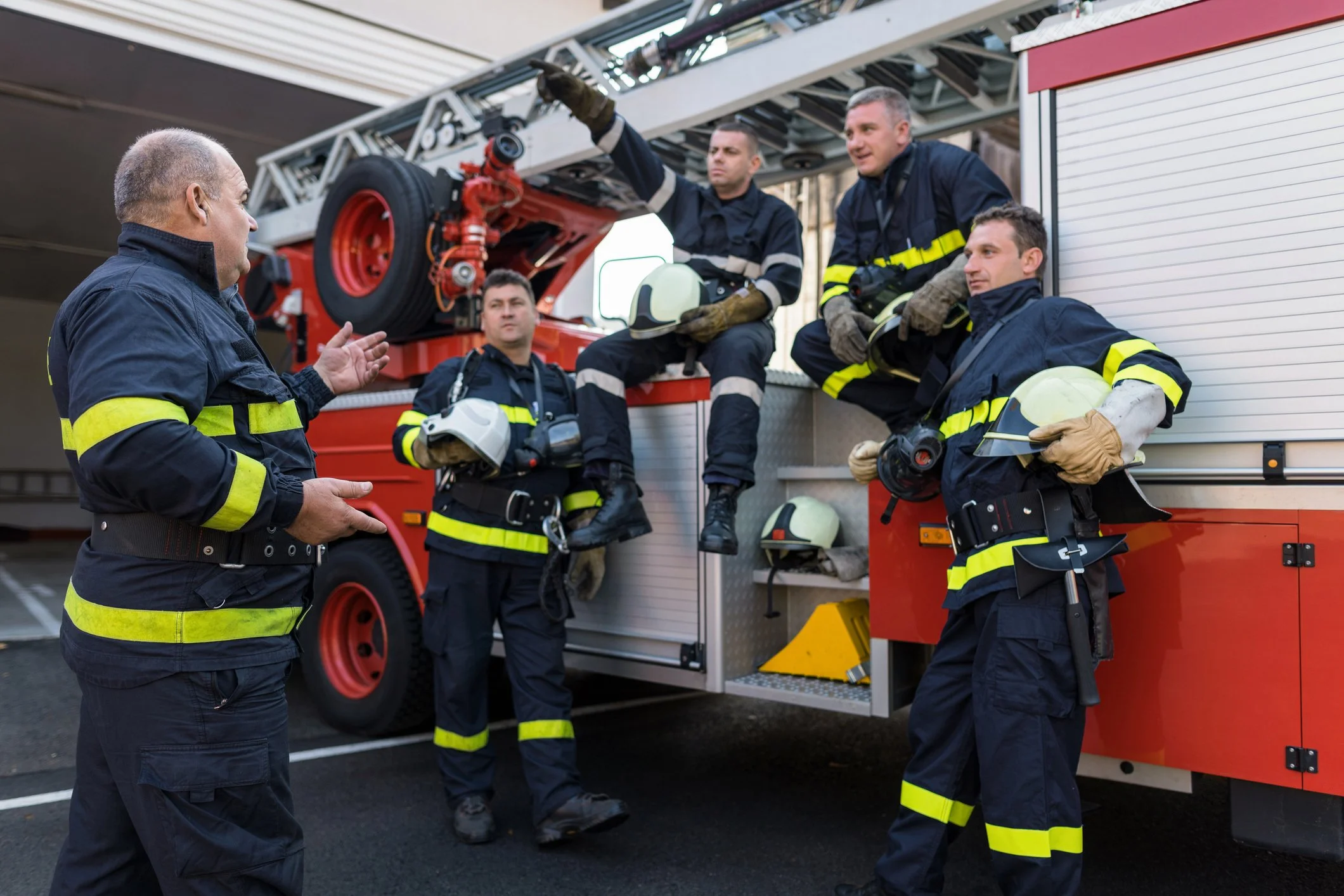 Group of firefighters in uniform taking a break and discussing near their fire truck, with rescue equipment and helmets visible.