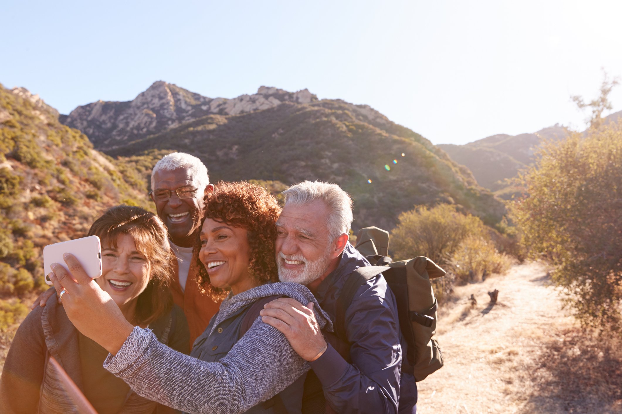 A group of five diverse older adults taking a selfie together outdoors in a mountainous area with bright sunlight.