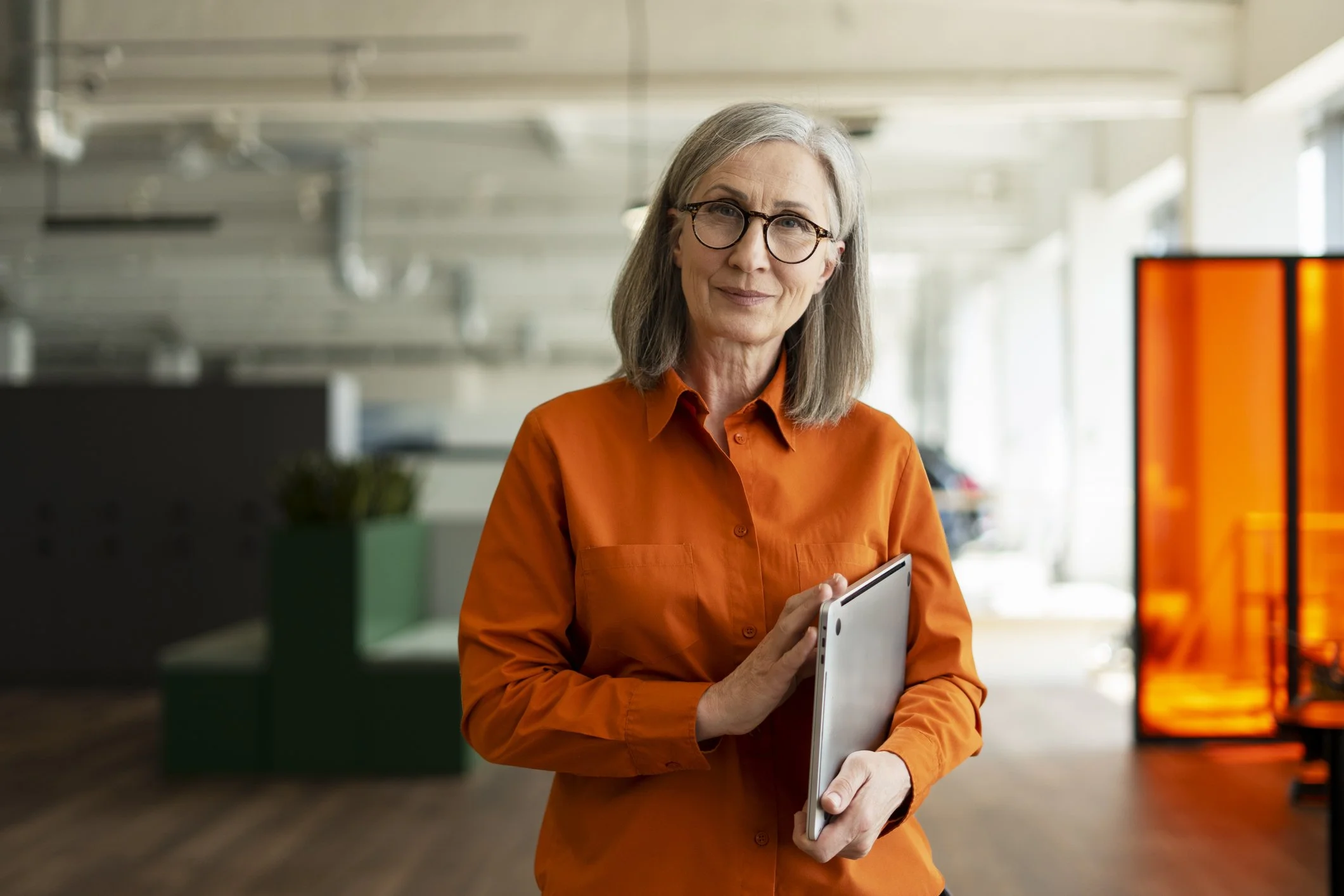 An elderly woman with gray hair, glasses, and an orange shirt holds a tablet in her hands. She is in an indoor setting with natural light coming through the windows and a brightly lit digital display to her right.