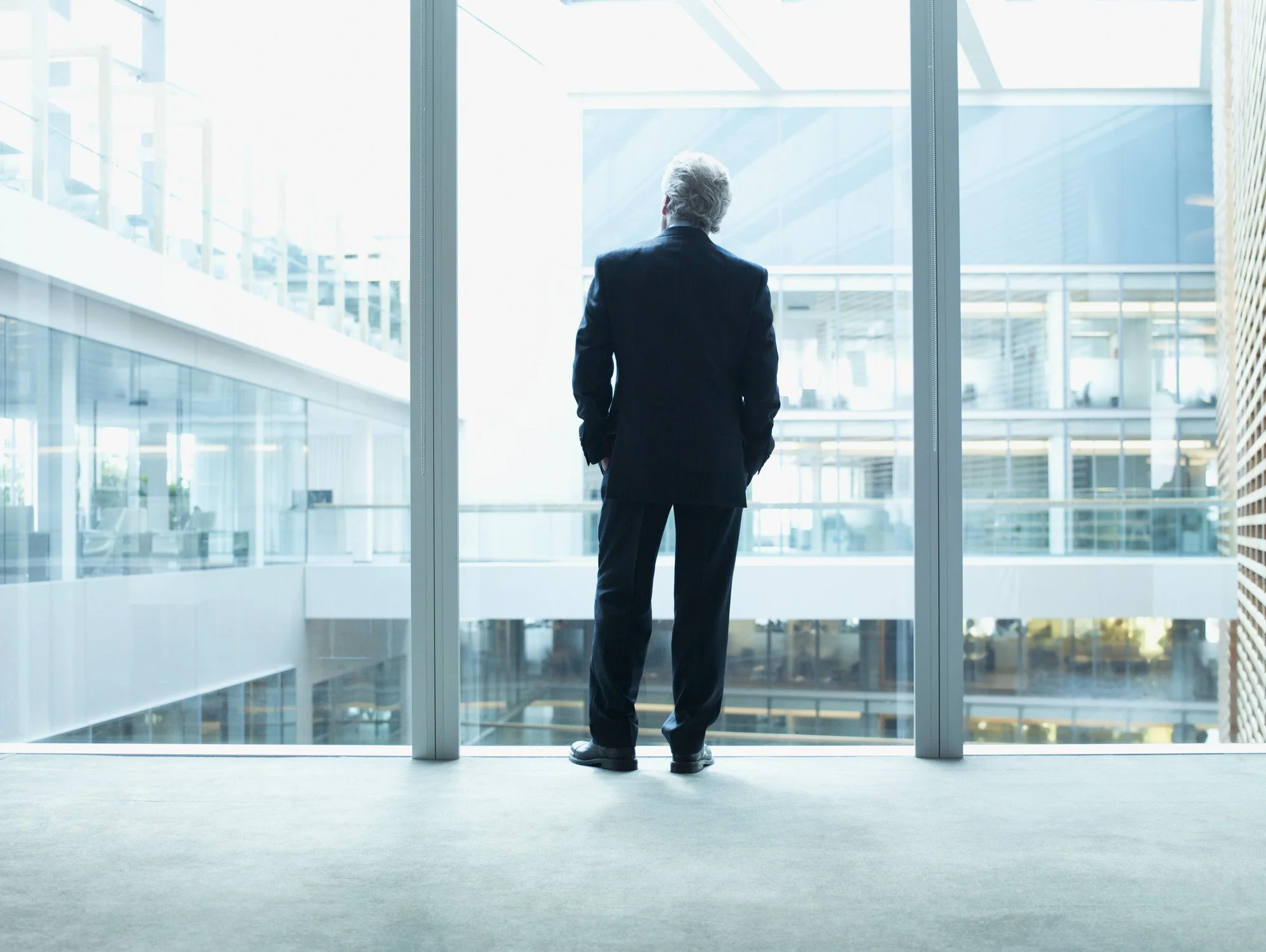 A man in a business suit looking out a large glass window in a modern office building