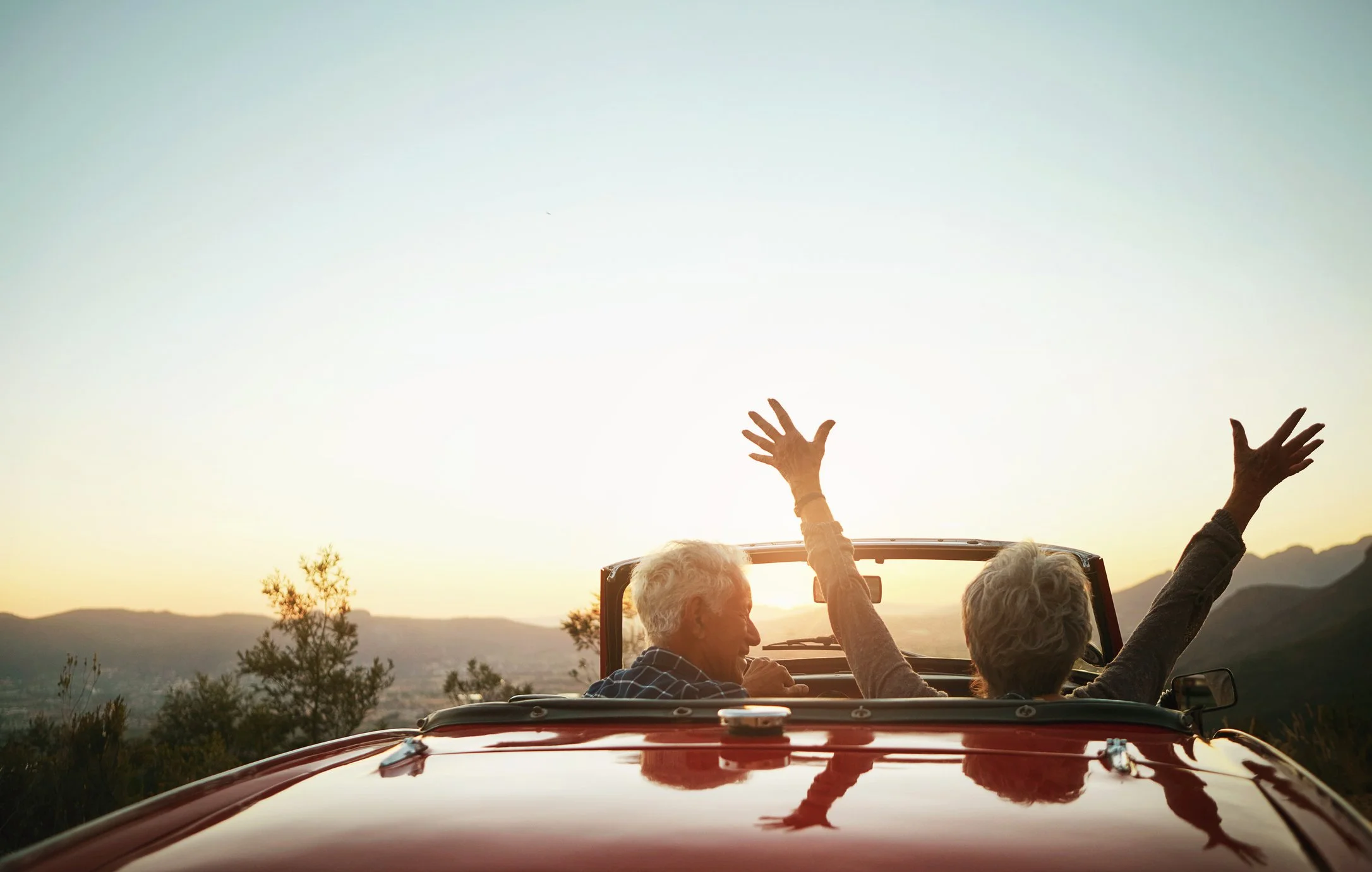 Two elderly people riding in a red convertible car at sunset, with one raising their hands in the air as they enjoy the scenic view of mountains and trees.