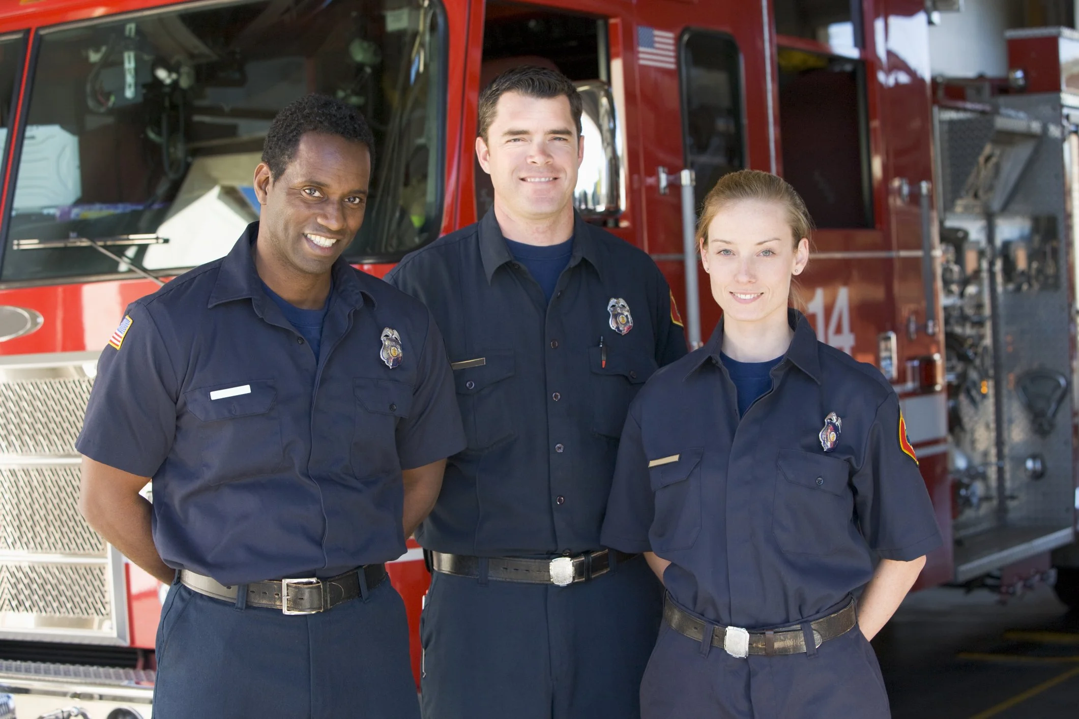 Three firefighters in uniform standing in front of a red fire truck, smiling at the camera.