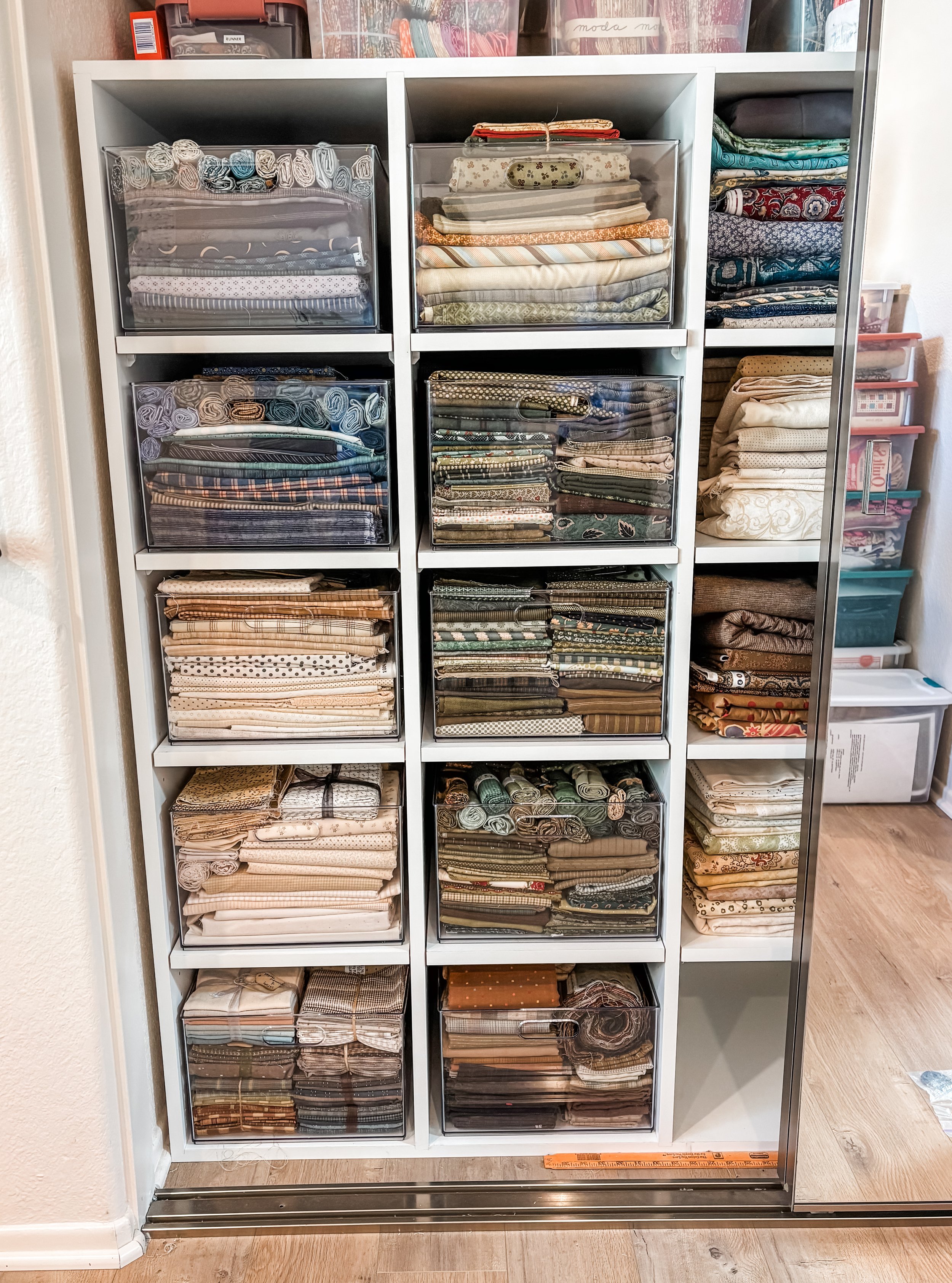 White shelving unit containing neatly folded and organized fabric and textiles, some protected in plastic covers, with additional storage boxes visible in the background.