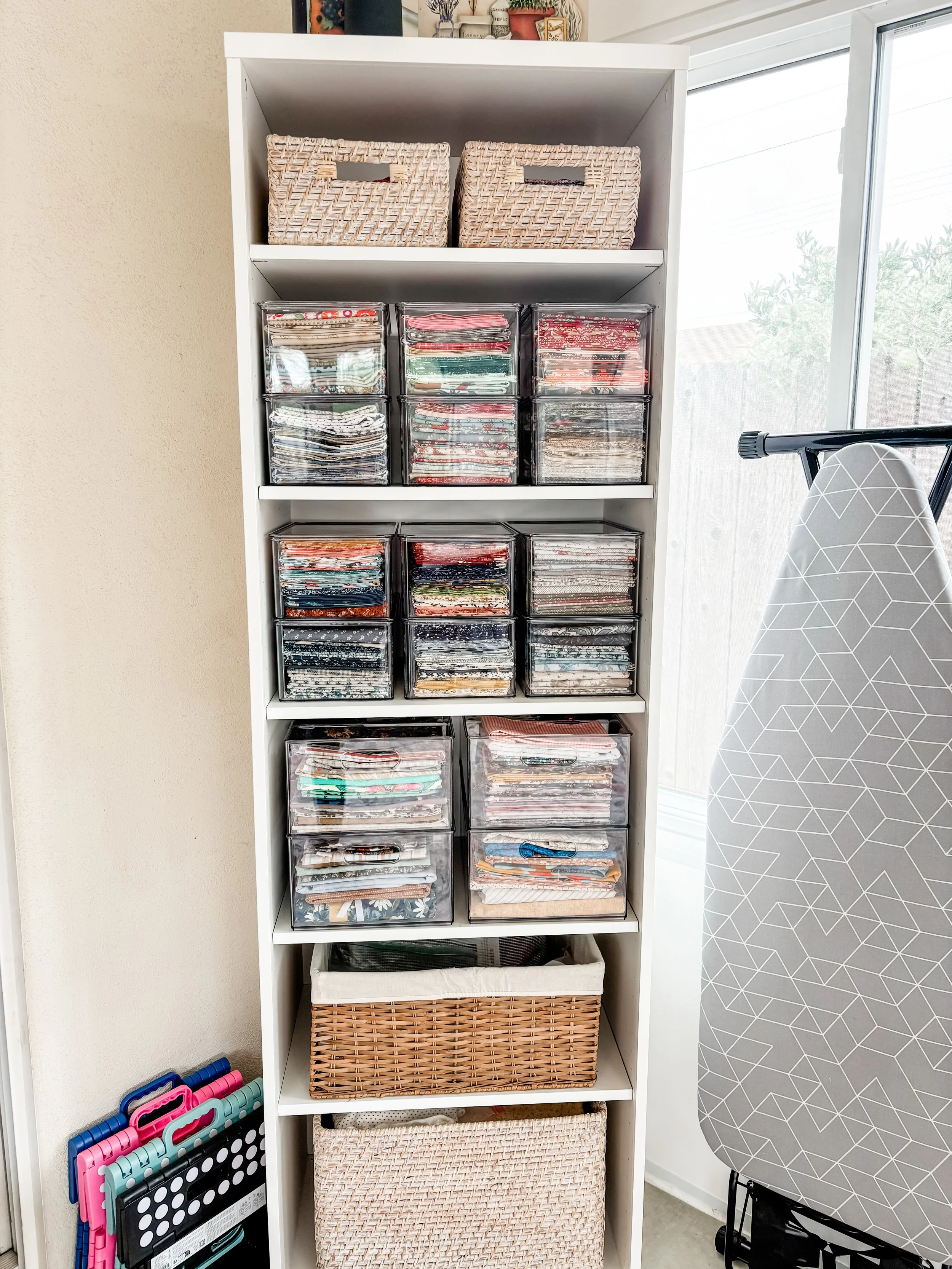A white shelf containing fabric scraps organized in clear plastic bins, with woven baskets on top and bottom, and ironing boards beside it.