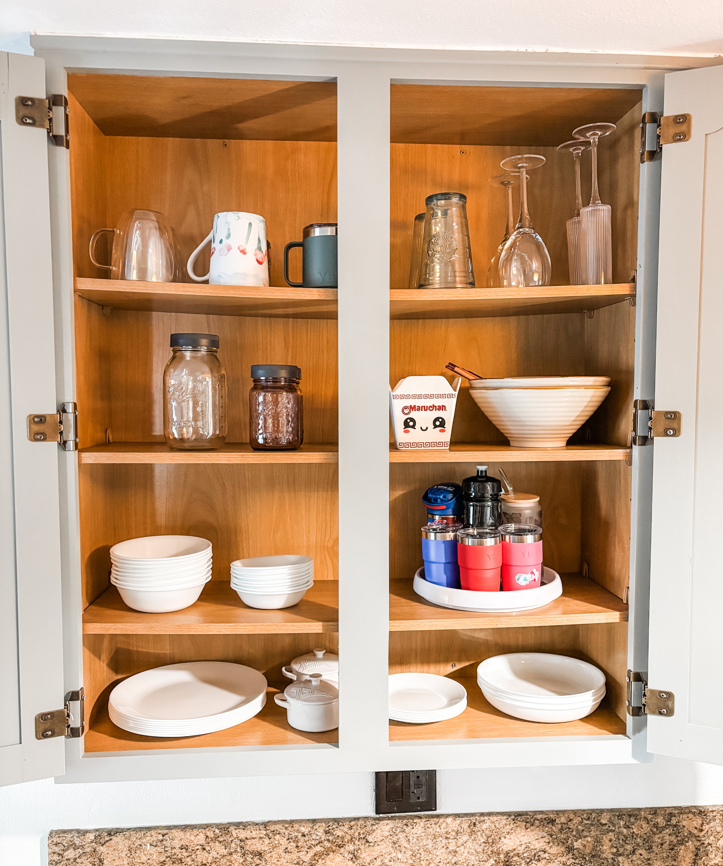 Open kitchen cabinet with glassware, bowls, and cups on wooden shelves.