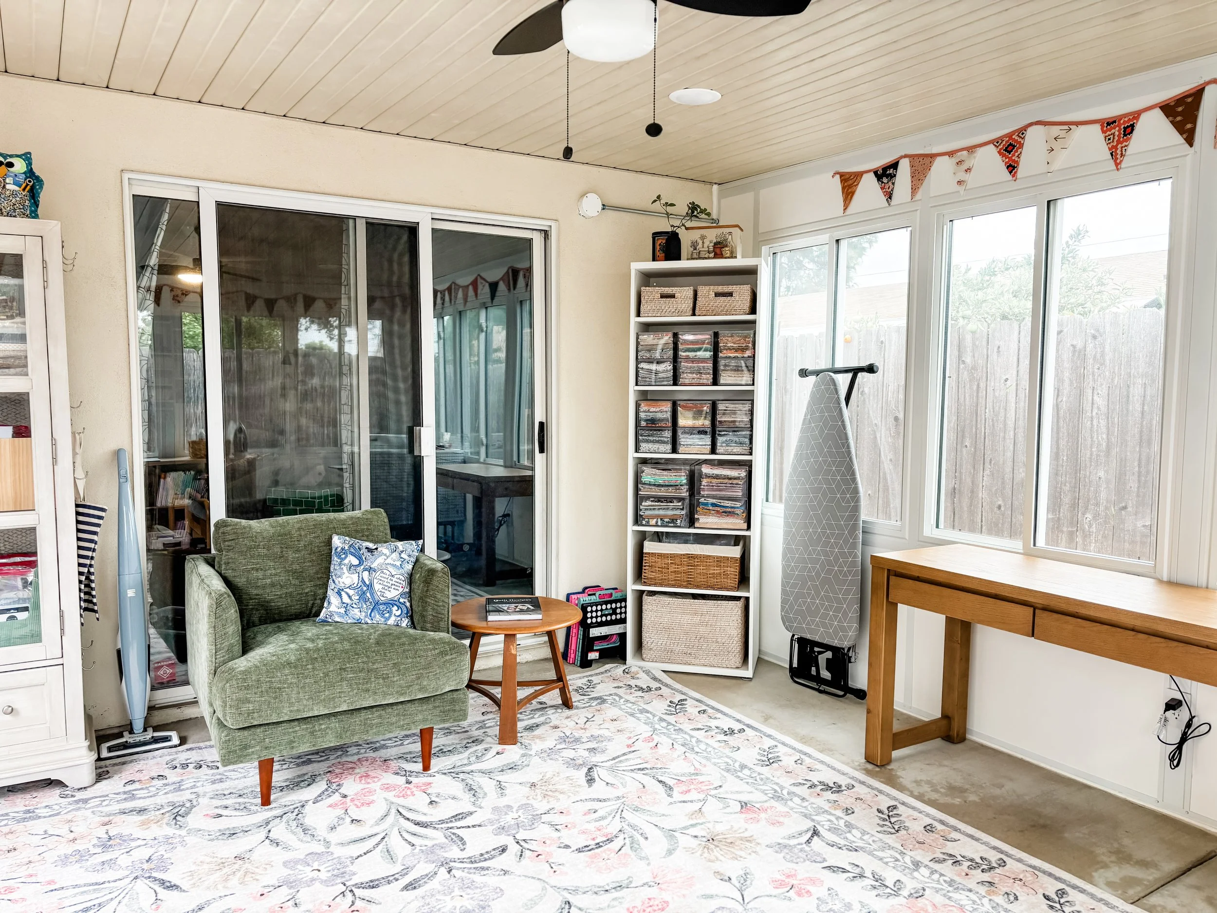 Sunroom with green armchair, light wood side table, windowed walls, entertainment center with magazines, ironing board, and wooden work table.