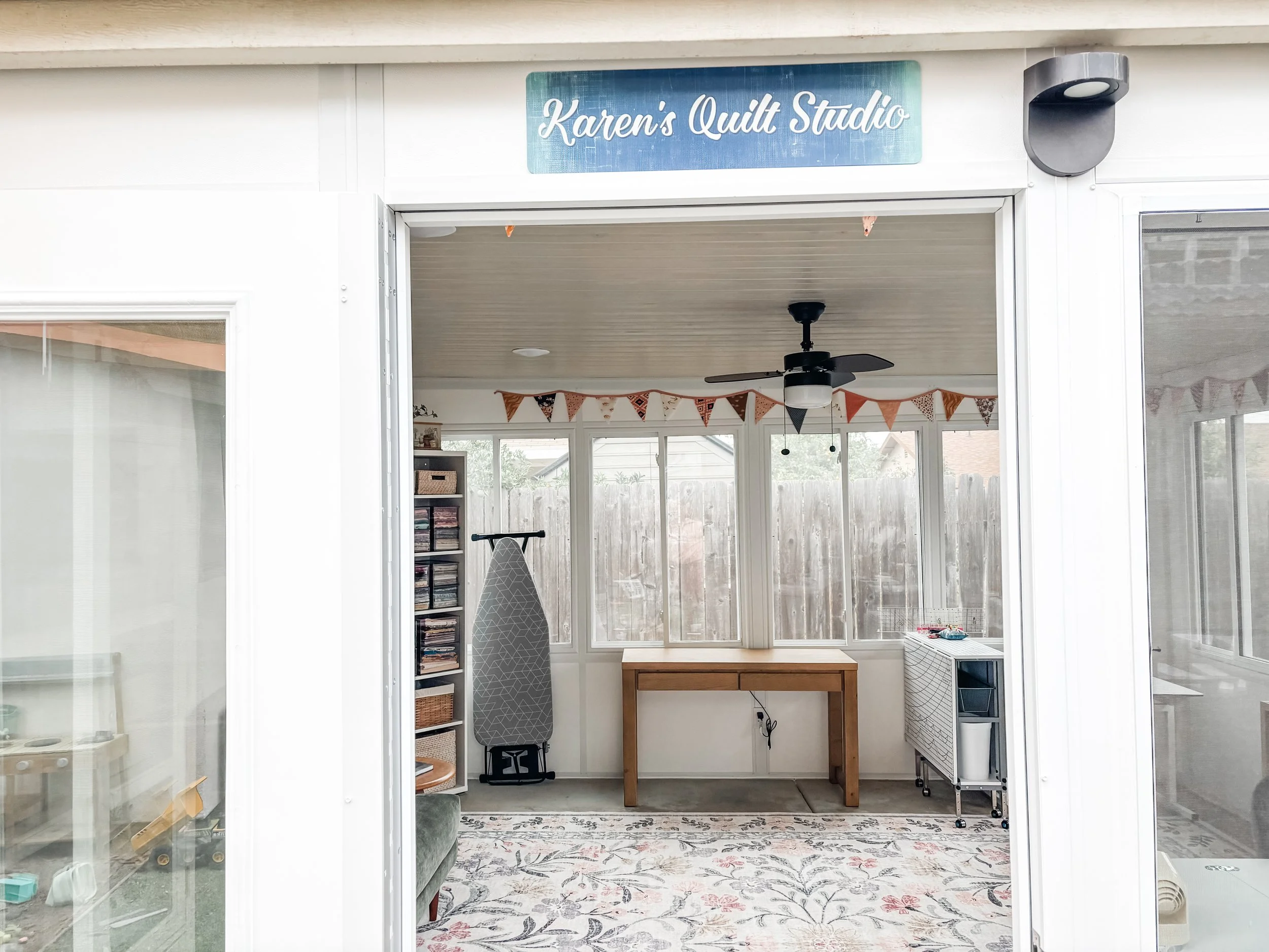 View through window showing quilt studio room with banners, ceiling fan, ironing board, bookshelf, and small storage unit.