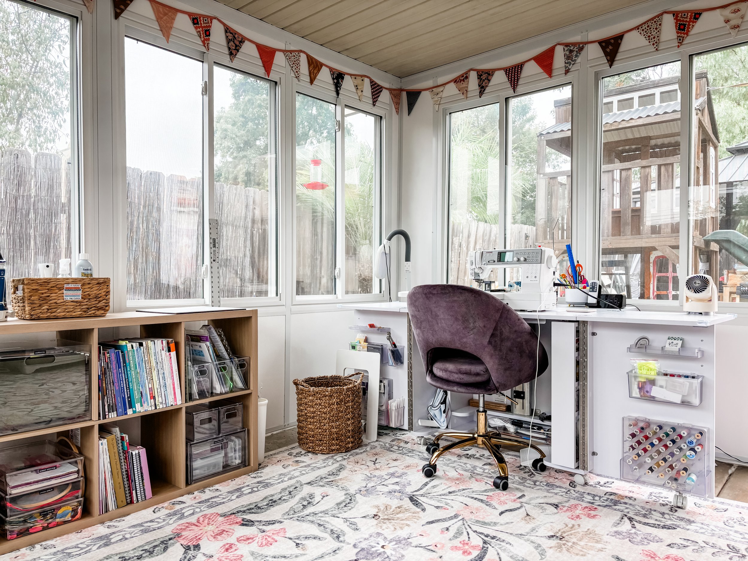 A bright craft or sewing room with large windows and a view outside, decorated with colorful bunting. The room has a bookshelf with books and storage boxes, a woven basket, a white desk with sewing and crafting supplies, and an office chair. There's 