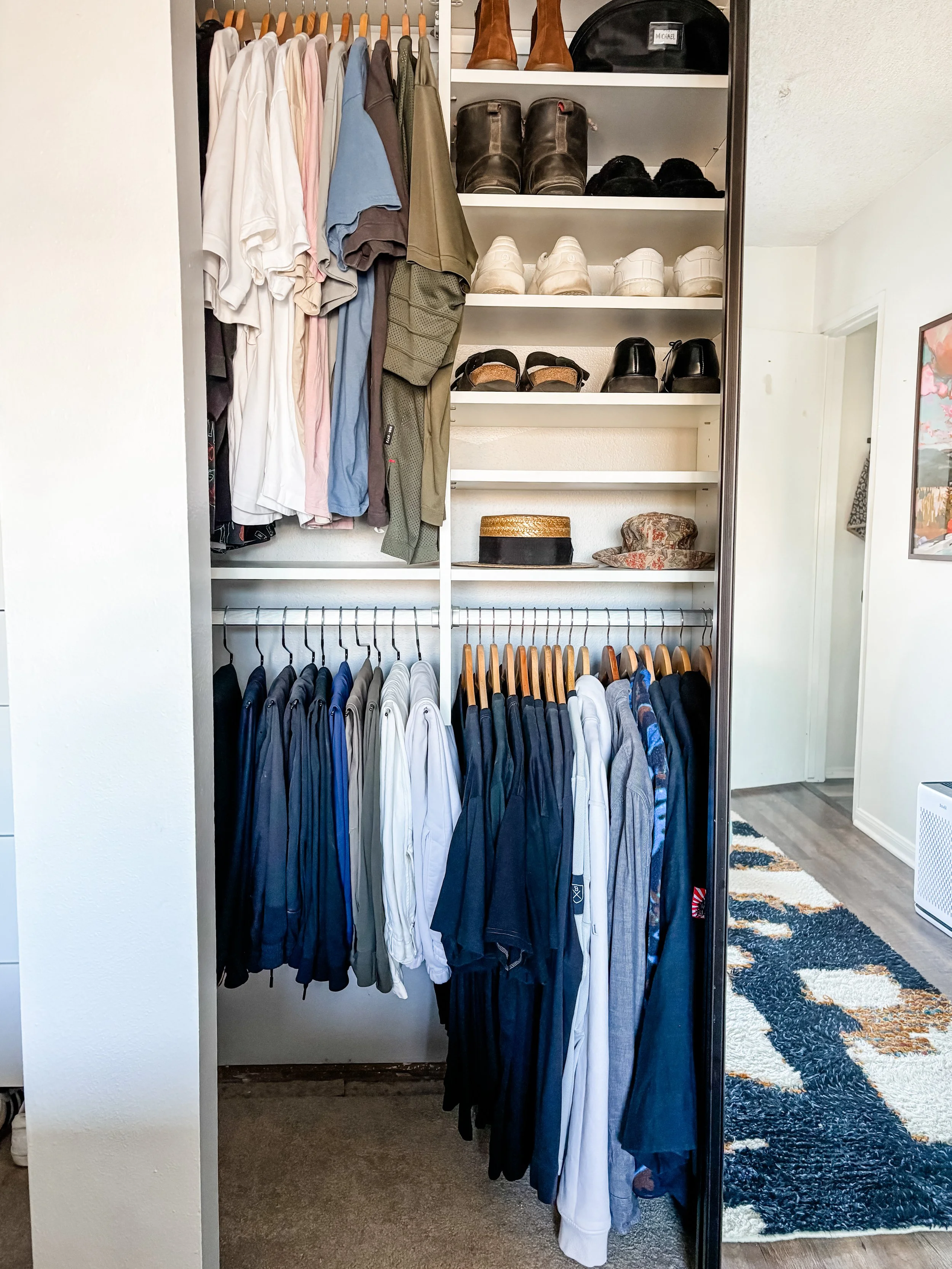 A closet with hanging clothes, shelves with shoes and accessories, and a patterned rug visible in a nearby room.