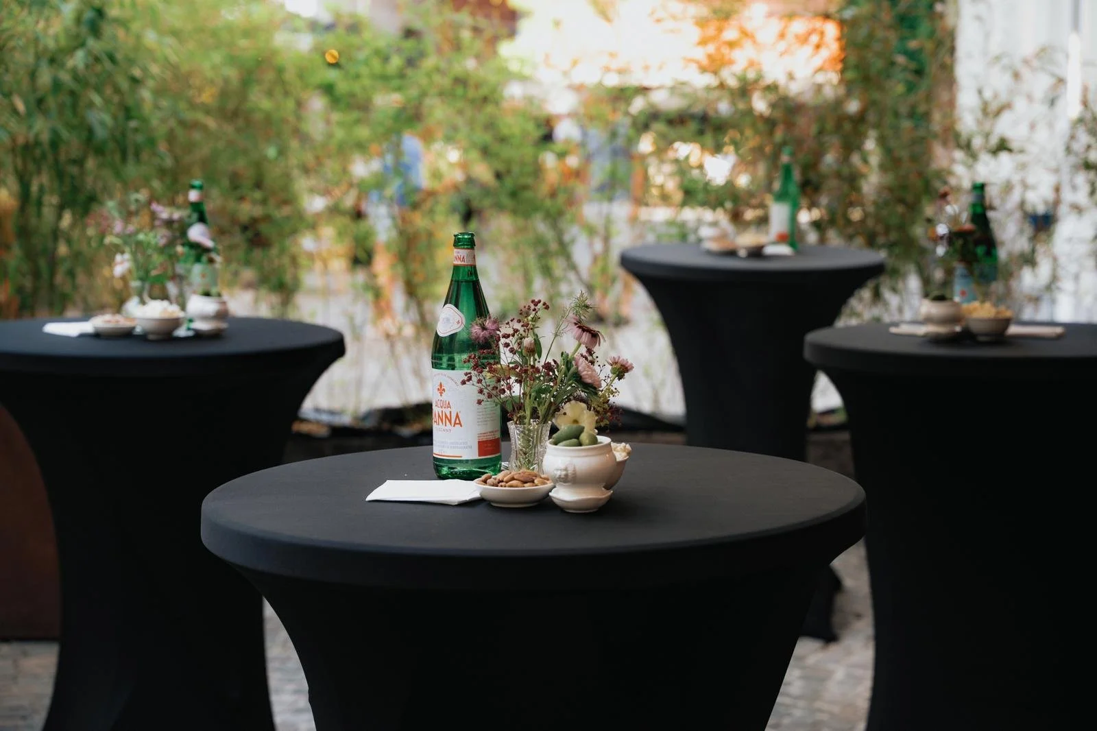 Outdoor event setup with black cocktail tables, floral centerpieces, bottles of water, small bowls of snacks, and a blurred background of trees and people.