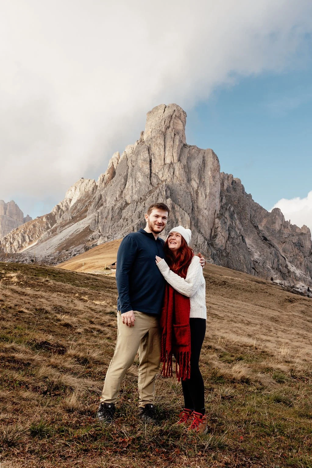 A couple standing close together smiling in front of a large mountain with rugged rocks, grassy terrain, and a partly cloudy sky.
