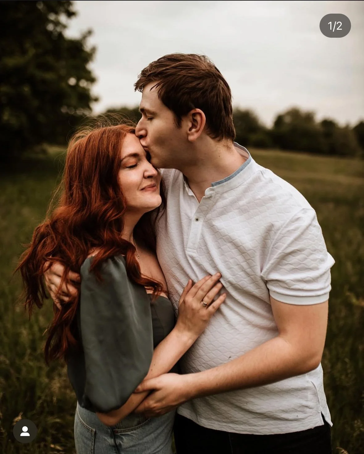 A man gives a gentle kiss on the forehead to a woman who is smiling with closed eyes, standing outdoors in a grassy area with trees in the background during overcast weather.
