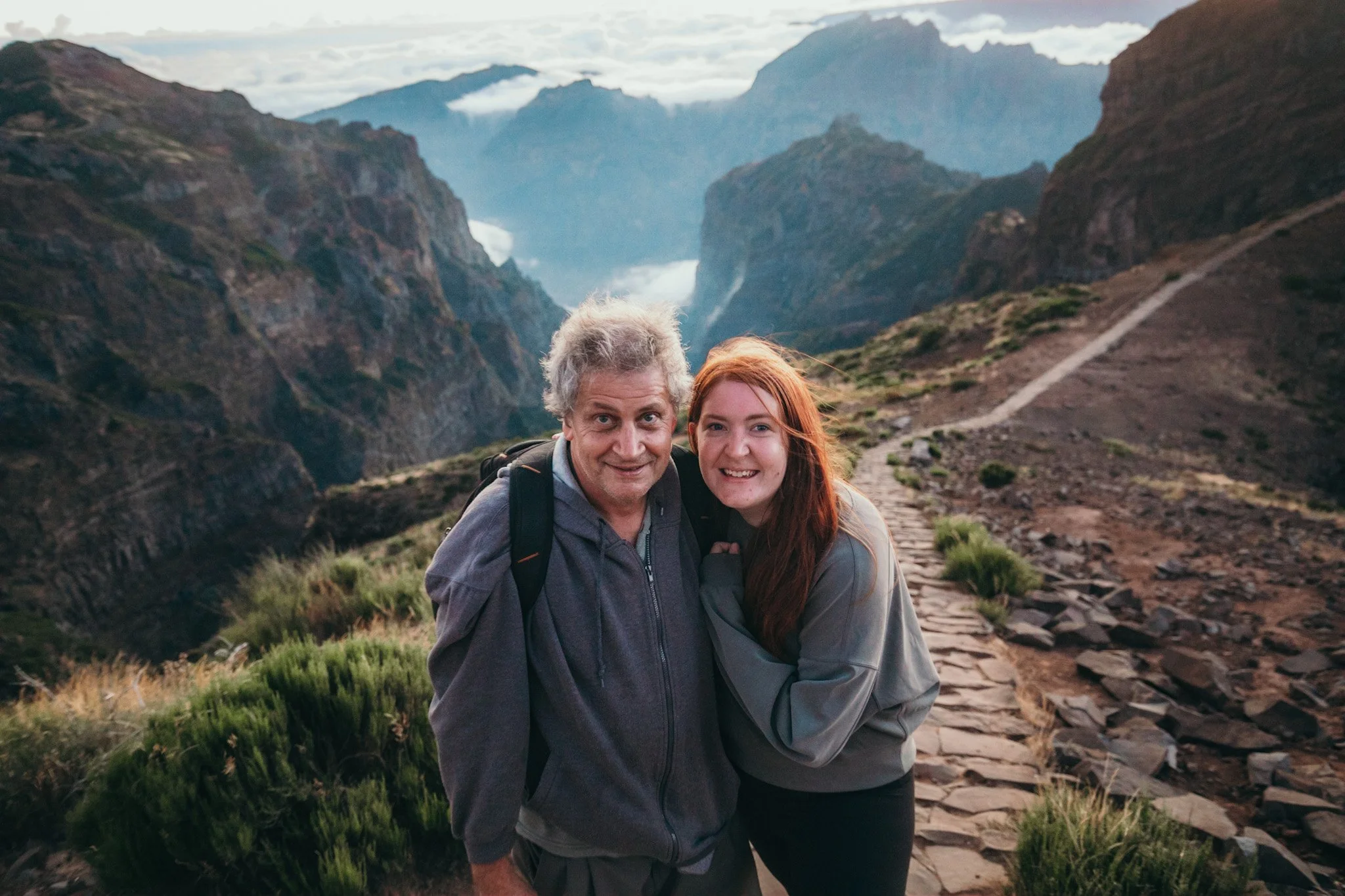 A man and a woman smiling and hugging on a mountainous hiking trail.