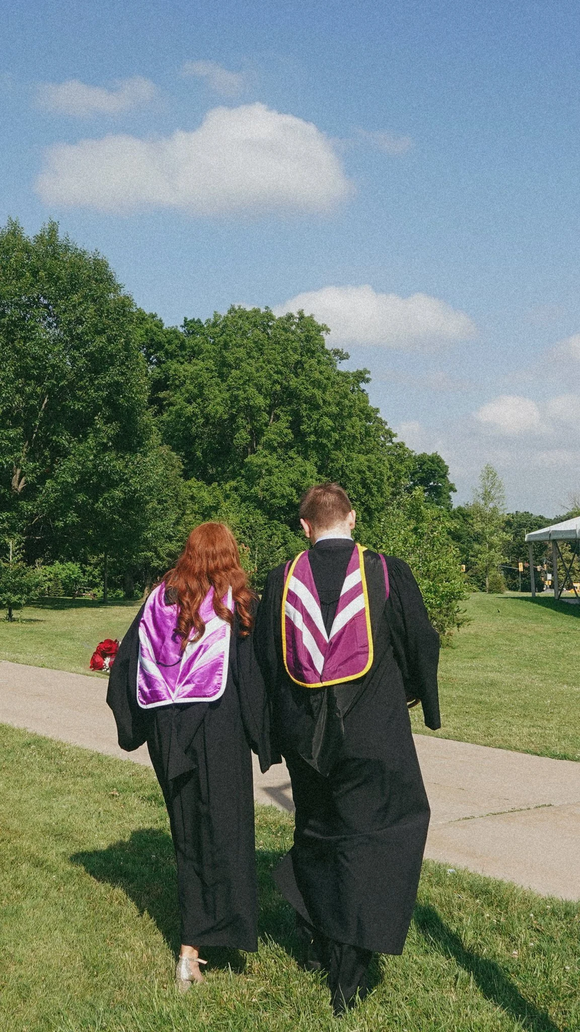 A man and woman in graduation gowns walking on a sidewalk outdoors, with green trees and a blue sky with clouds in the background.