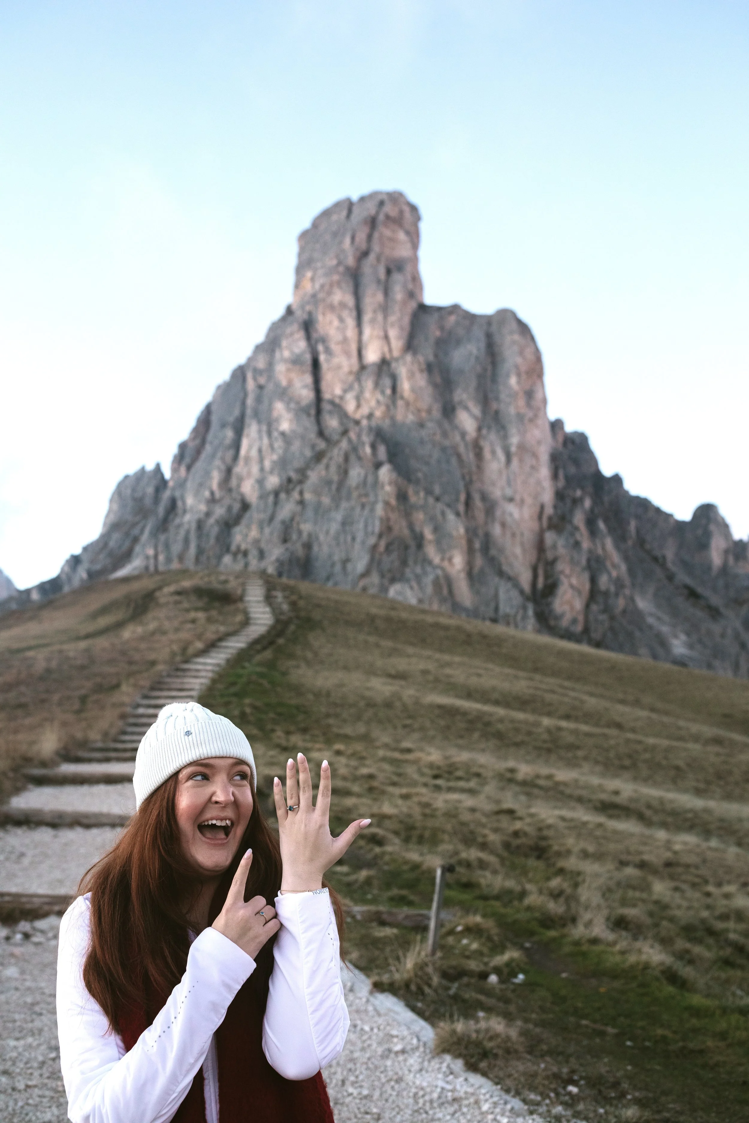A woman with long red hair wearing a white beanie and white jacket, smiling and pointing at her wedding ring, in front of a mountain landscape with a rocky mountain and a trail leading up the hill.