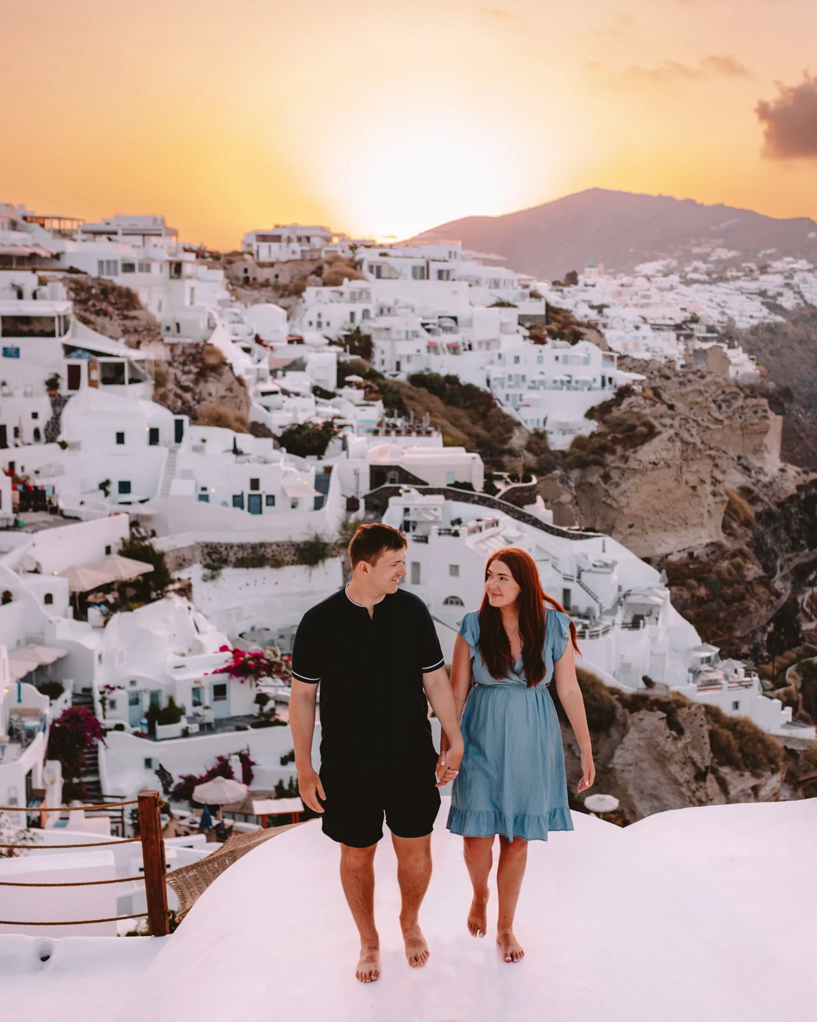 A couple walking hand in hand on a white rooftop at sunset in a Greek village with white buildings on a hillside.