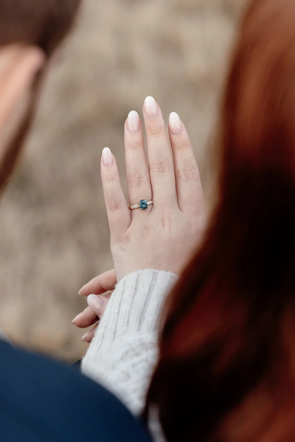 A woman with red hair wears a white sweater and displays her left hand with a turquoise gemstone ring on her ring finger, against a blurred neutral background.