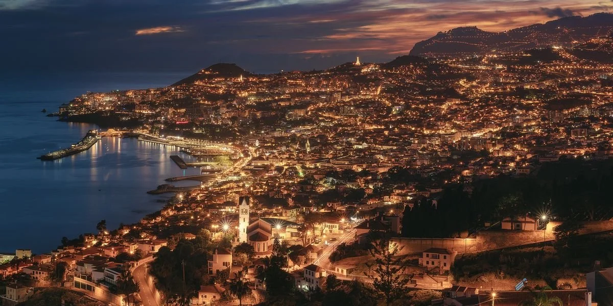 A cityscape of a coastal city at dusk with numerous lights, a harbor, and hills in the background.