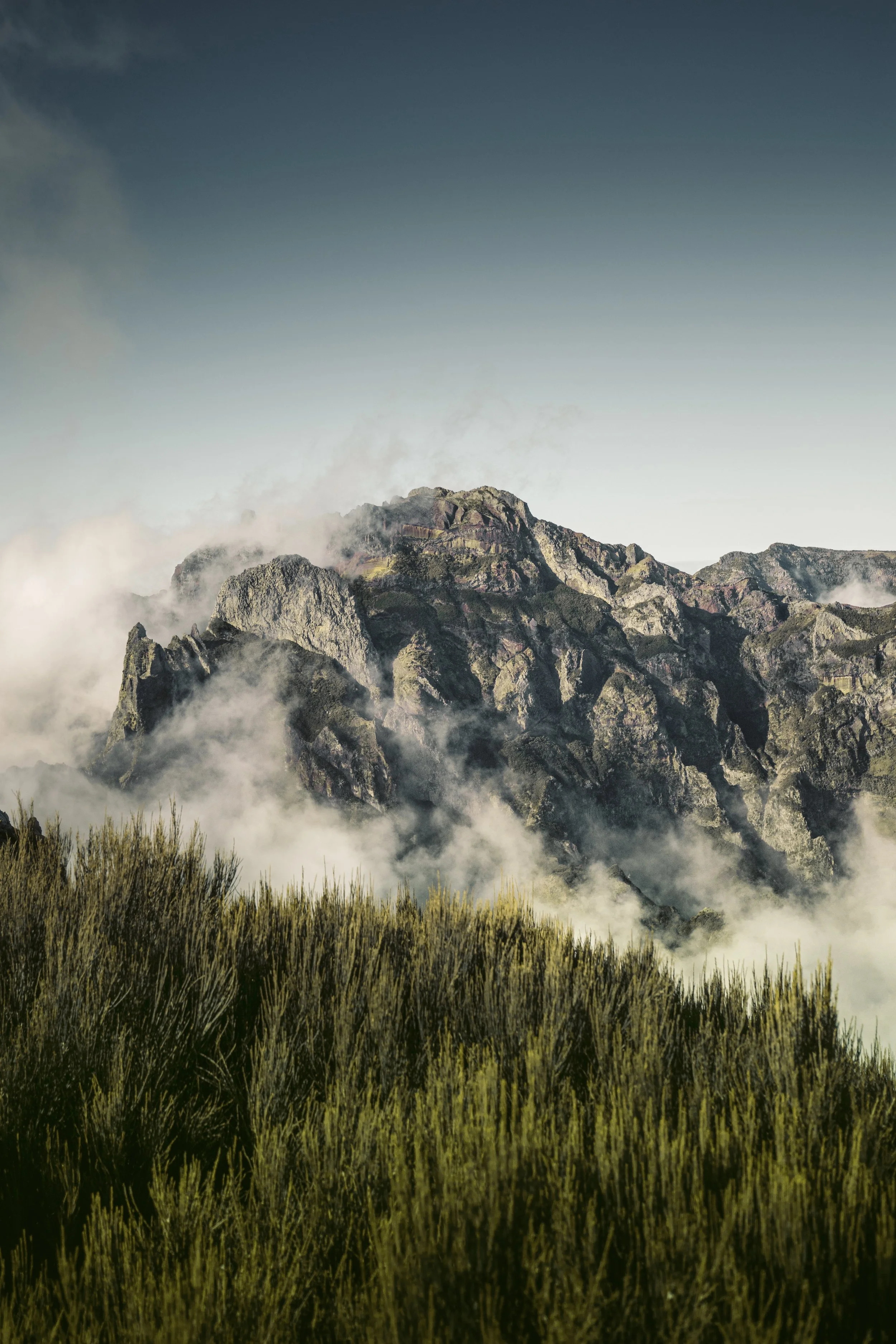 A mountainous landscape with rocky peaks partially covered by clouds and fog, and green vegetation in the foreground.