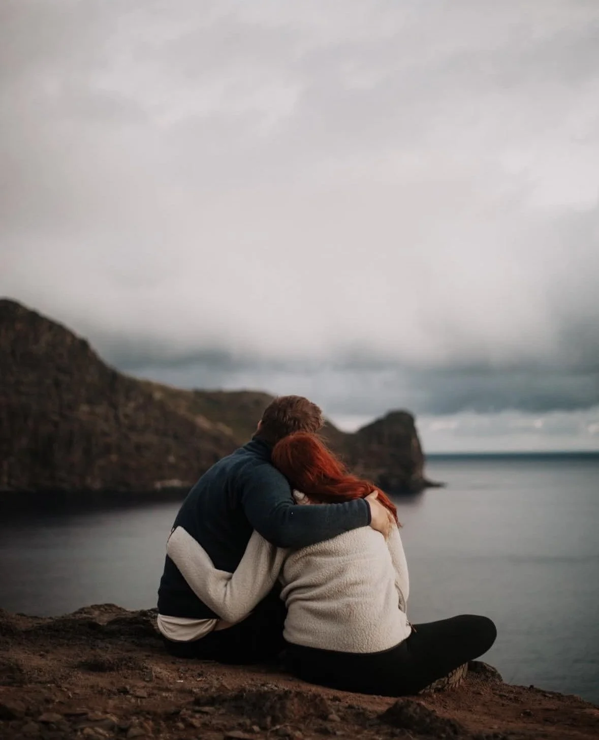 A couple sitting on a rocky ledge by the water, embracing and looking at the ocean under an overcast sky.