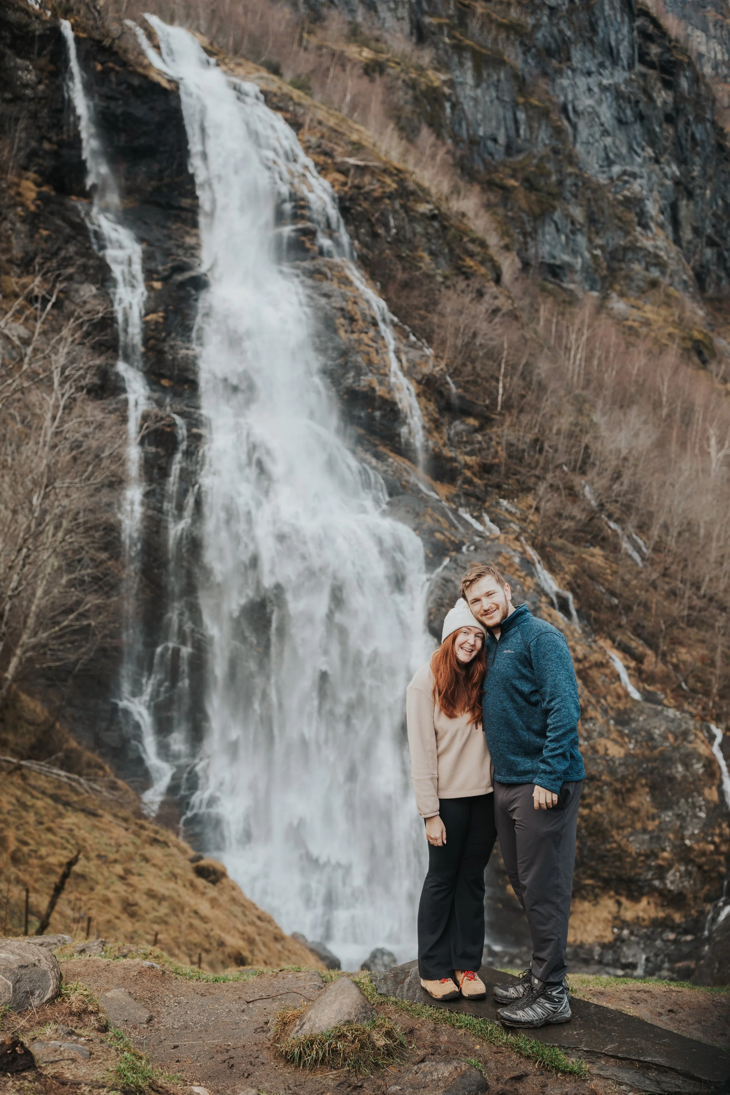 A couple standing together smiling in front of a large waterfall on a rocky outdoor trail.