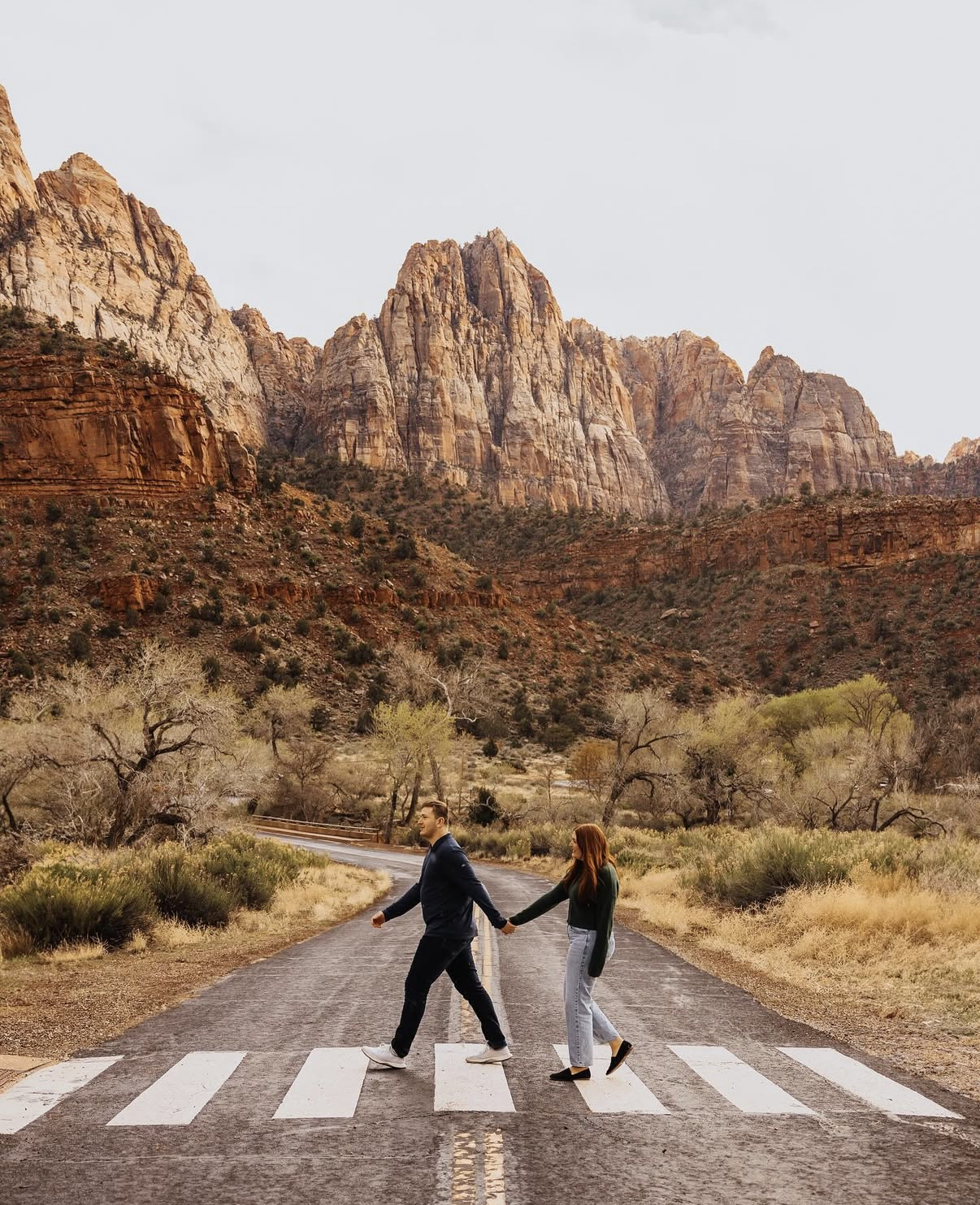 A couple holding hands and walking across a crosswalk on a deserted road with a mountainous desert landscape in the background.