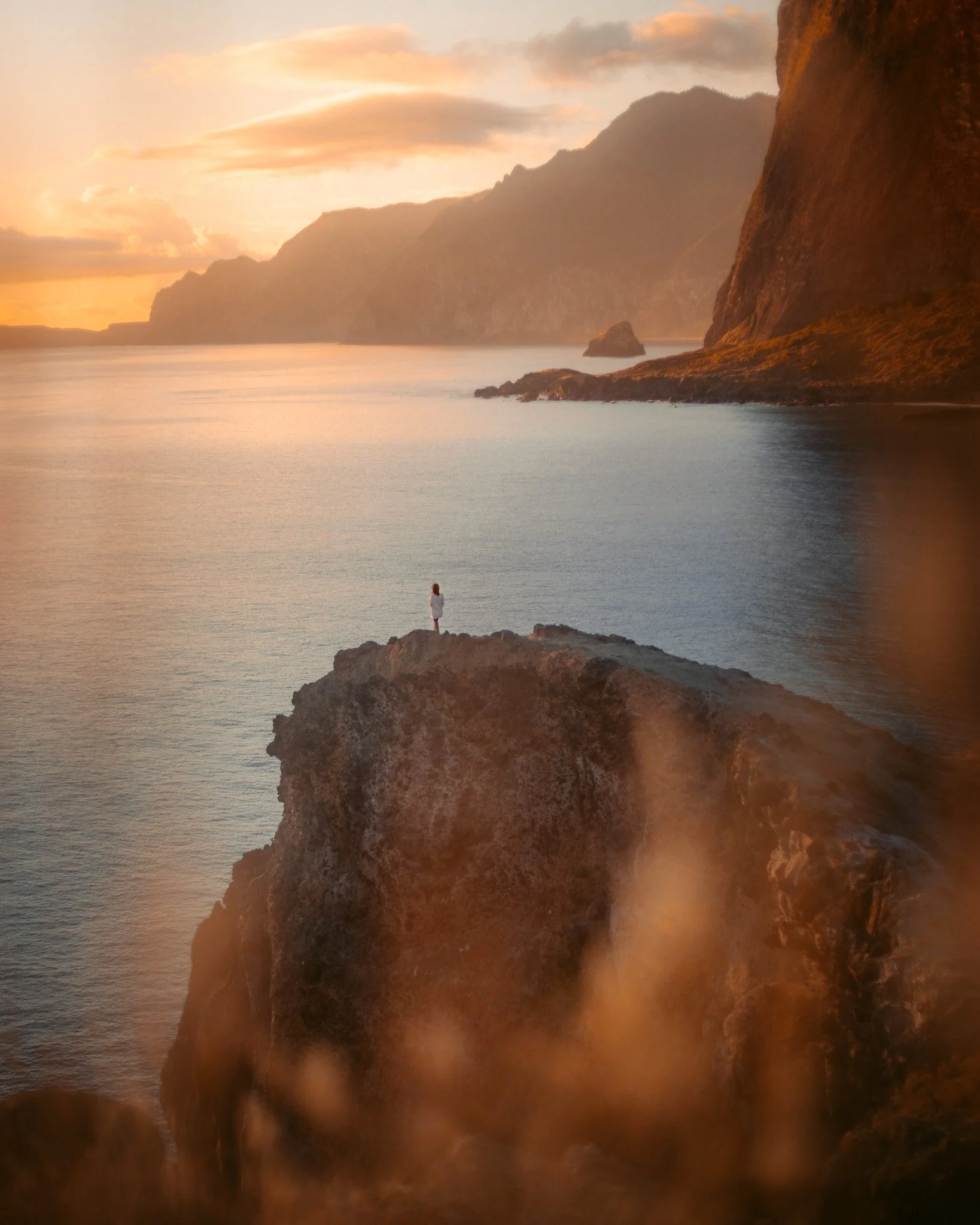 A person standing on a rocky cliff overlooking a calm body of water at sunset, with mountains in the background and a warm, golden sky.