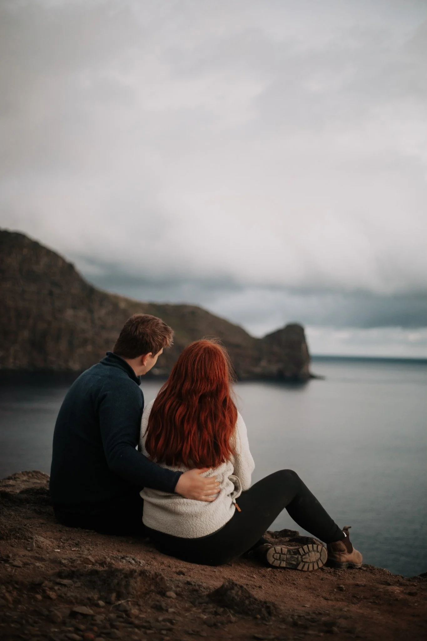 A couple sitting on a rocky cliff near the water, with their backs to the camera, looking out at a cloudy, mountainous landscape across a body of water.