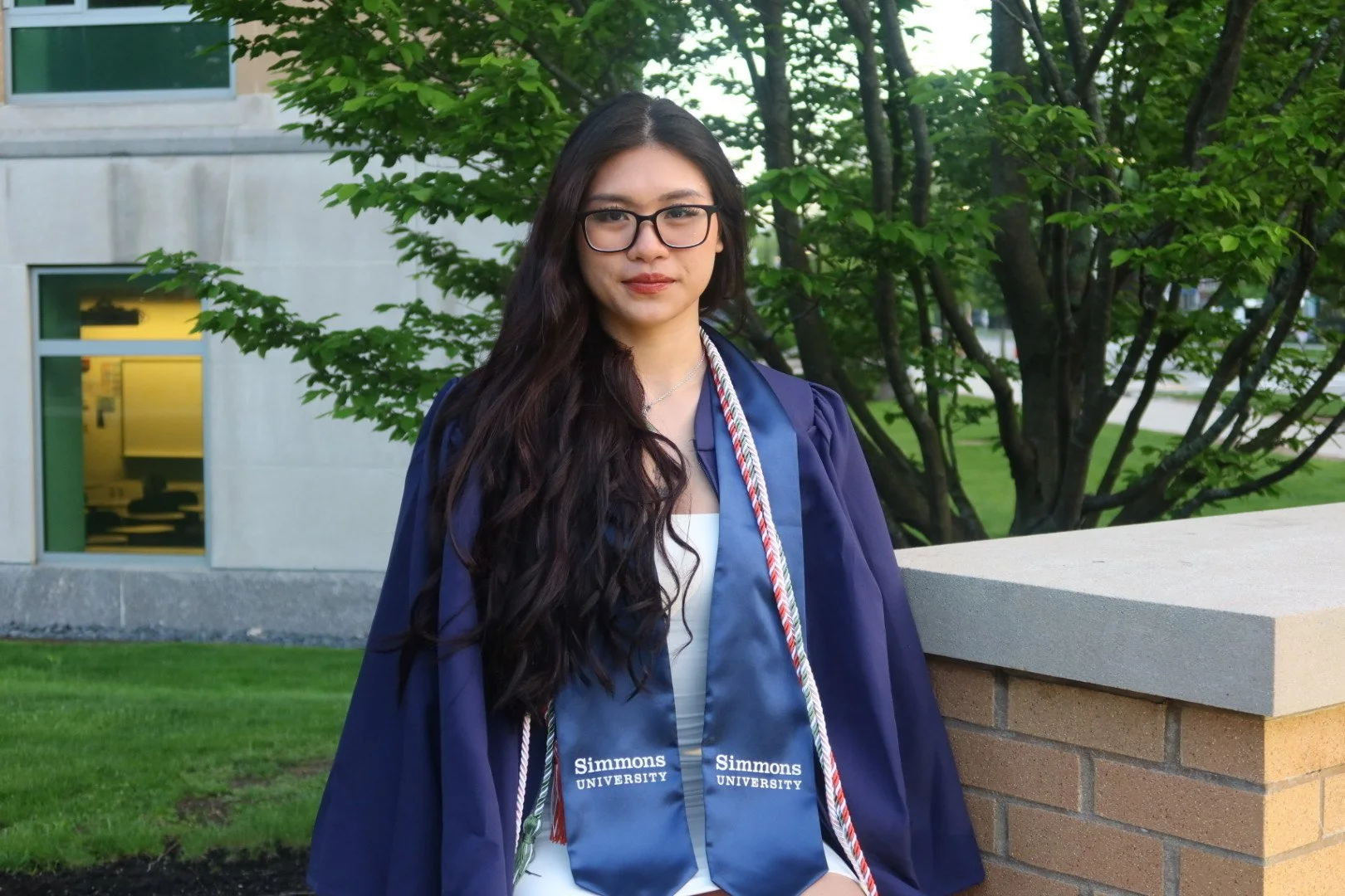 A young woman with long dark hair and glasses in a graduation gown and stole stands outdoors near a brick wall with green trees and a building in the background.
