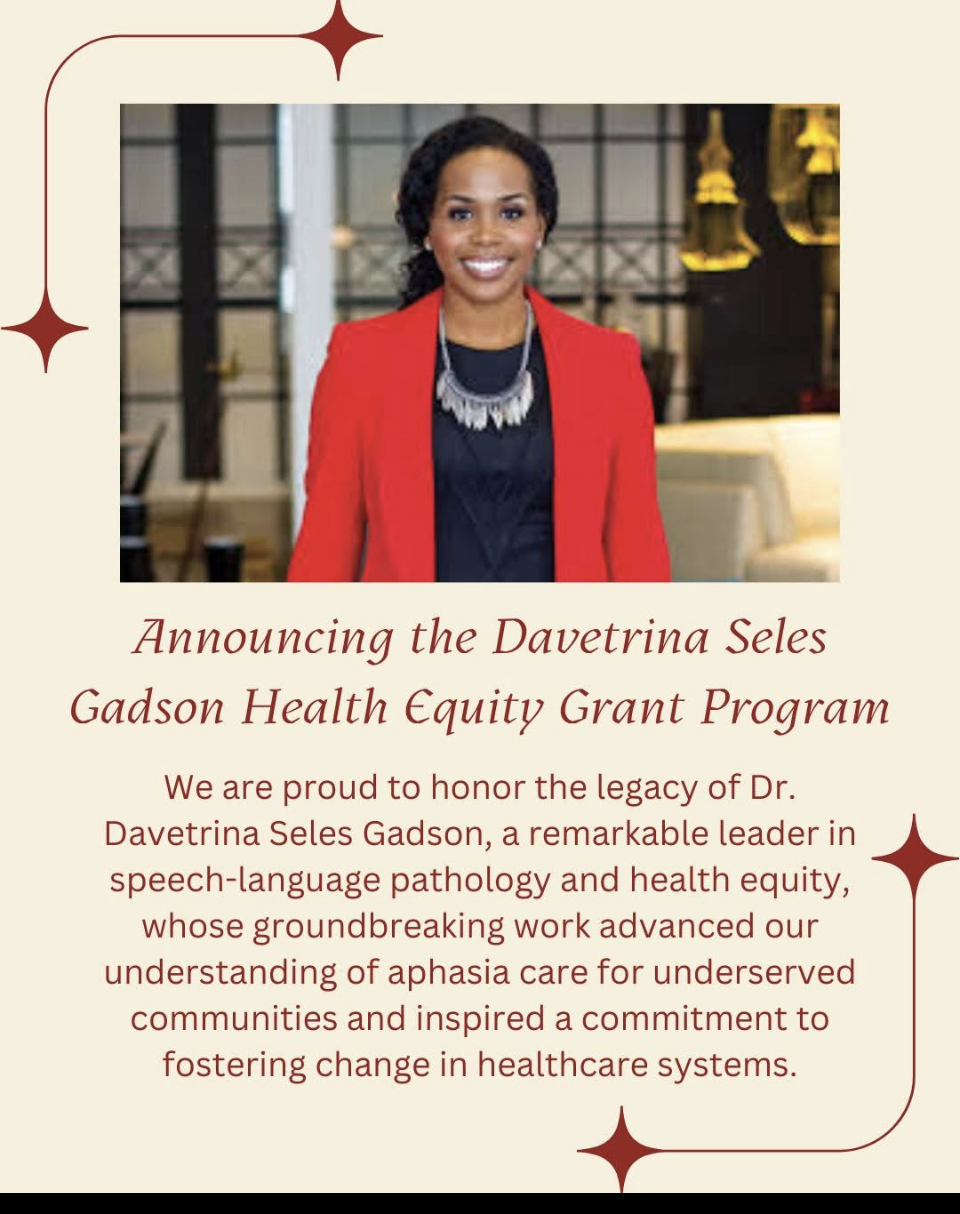 A woman with black curly hair and a bright smile, wearing a red blazer and statement necklace, standing inside a modern, warmly lit space with hanging lights and a cozy seating area.