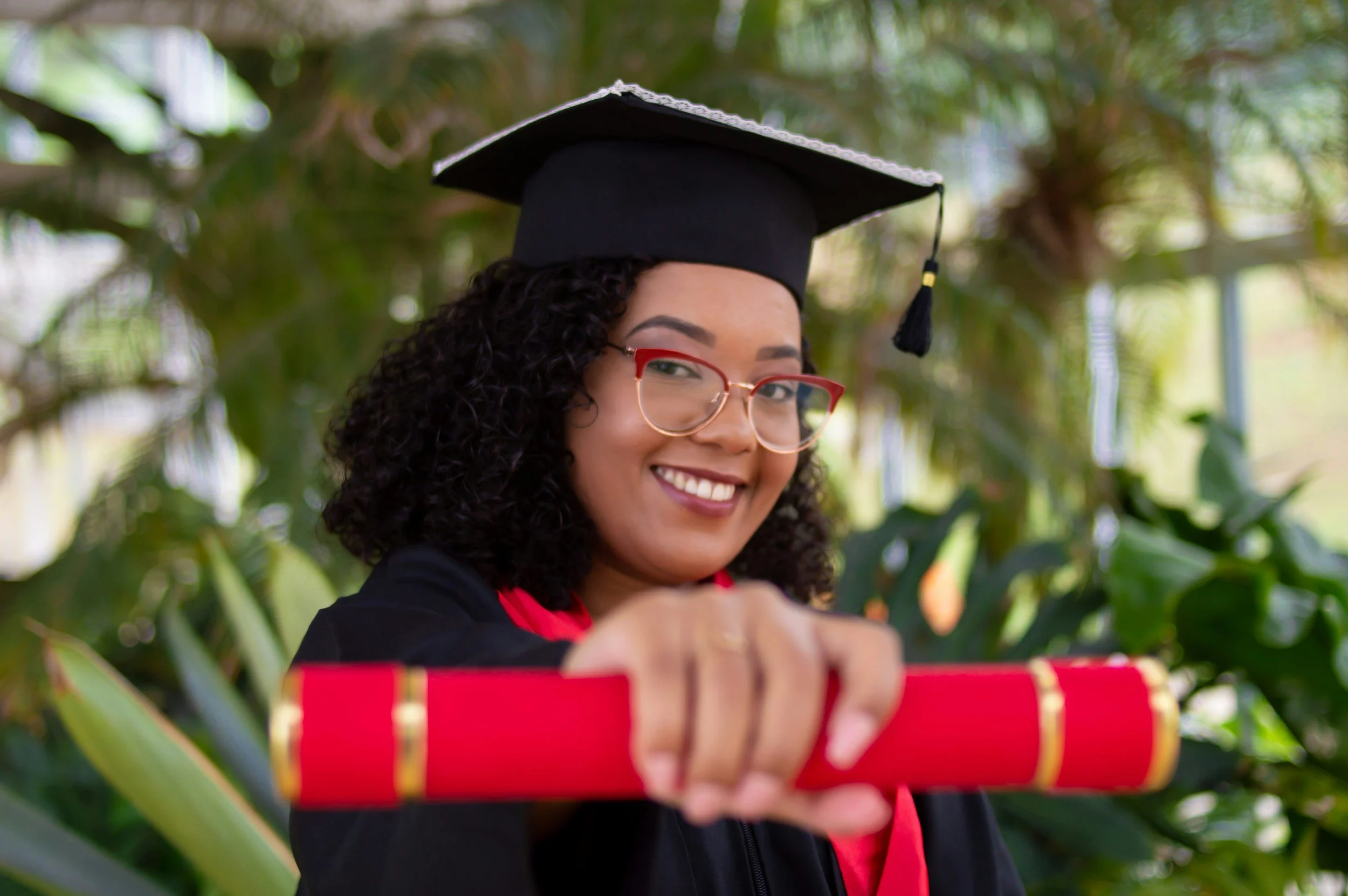 A young woman with curly hair and glasses wearing a graduation cap and gown, smiling and holding a rolled-up diploma in a lush, green indoor setting.